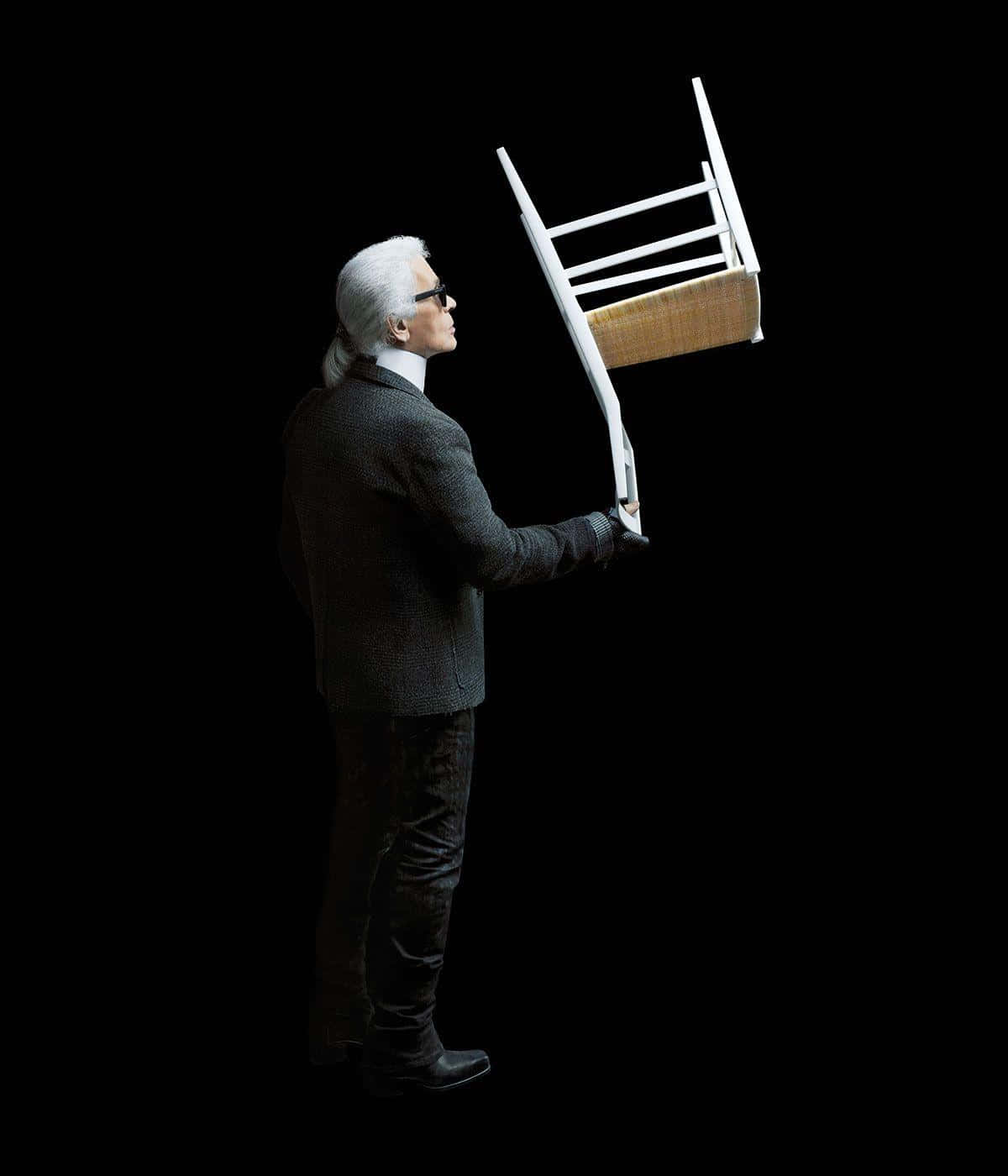 Karl Lagerfeld 桌布 壁紙 — 一名身穿深色西裝、戴著太陽眼鏡的男子，在黑色背景前倒舉著一張白色椅子。
