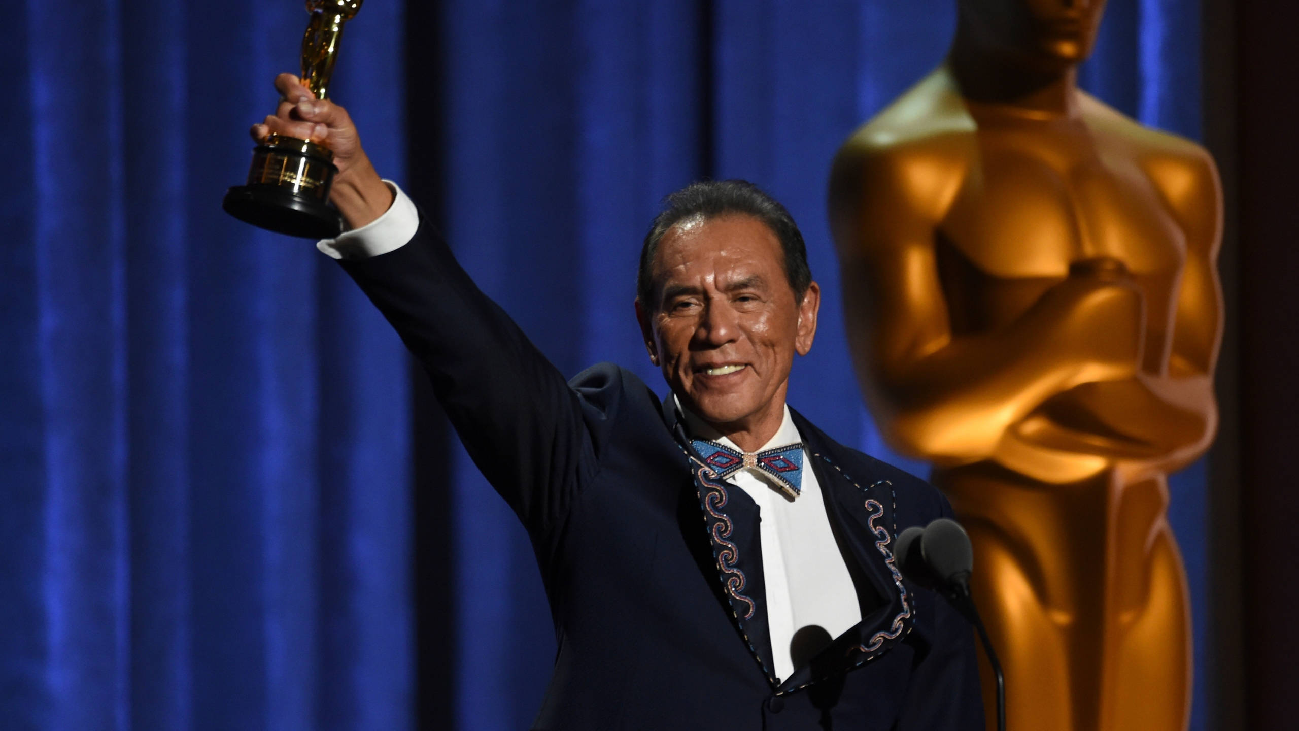 actor 桌布 壁紙 — 一名身穿深色西裝、繫著圖案領結的男子，在藍色帷幕舞台上得意地高舉奧斯卡獎座，旁邊有一座金色奧斯卡雕像。