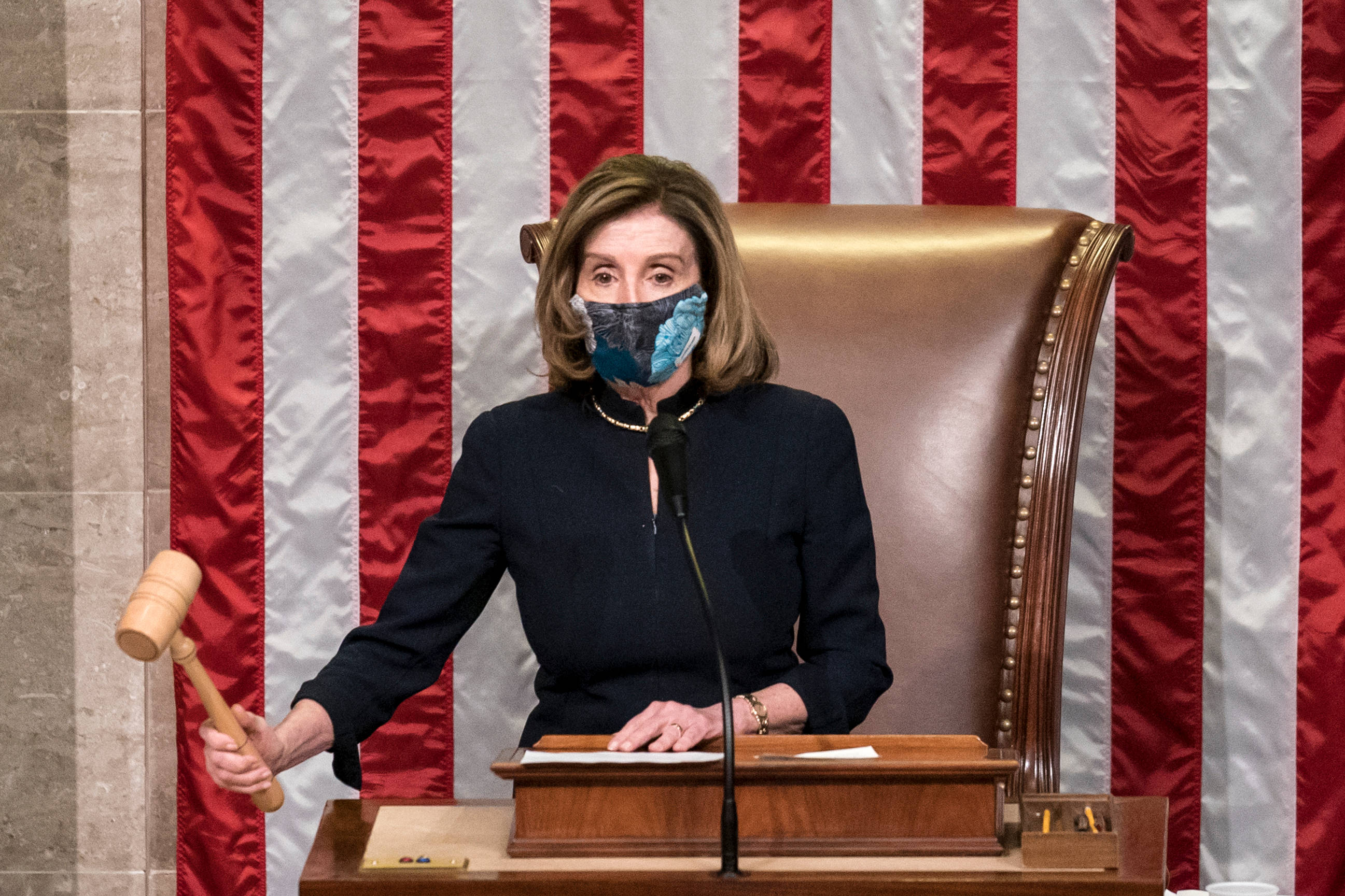 Nancy Pelosi 桌布 壁紙 — 一名身穿深色西裝及圖案口罩的女性坐在木桌前，在紅白條紋背景前手持木槌。