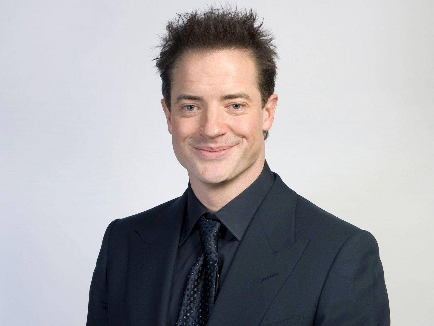 Celebrity 桌布 壁紙 — 一名身穿深色西裝及圖案領帶的男子，在純淺灰色背景前溫暖地微笑。