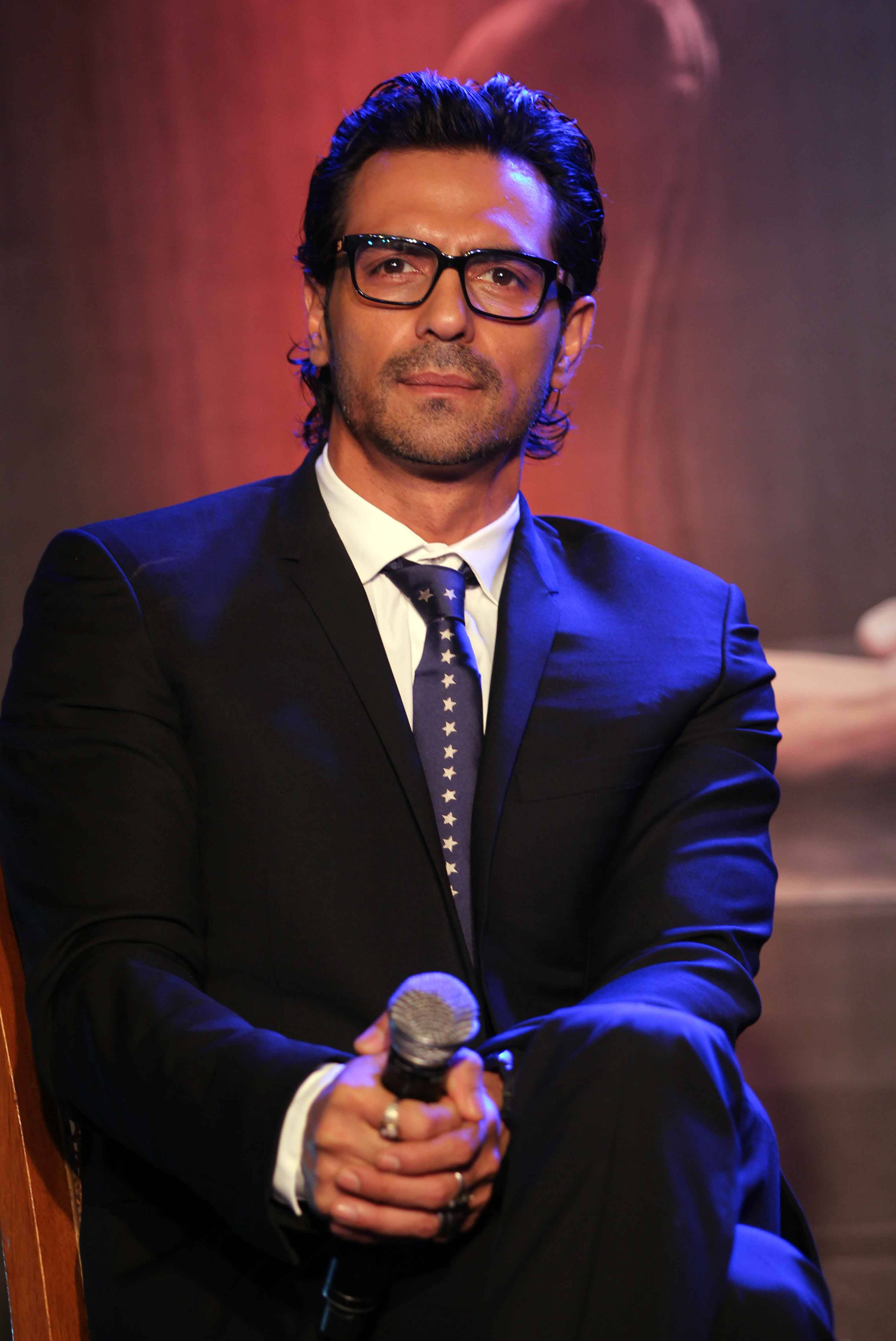 Arjun Rampal 桌布 壁紙 — 一名身穿深色西裝及星形圖案領帶的男子，在戲劇性的藍色舞台燈光下握著麥克風。