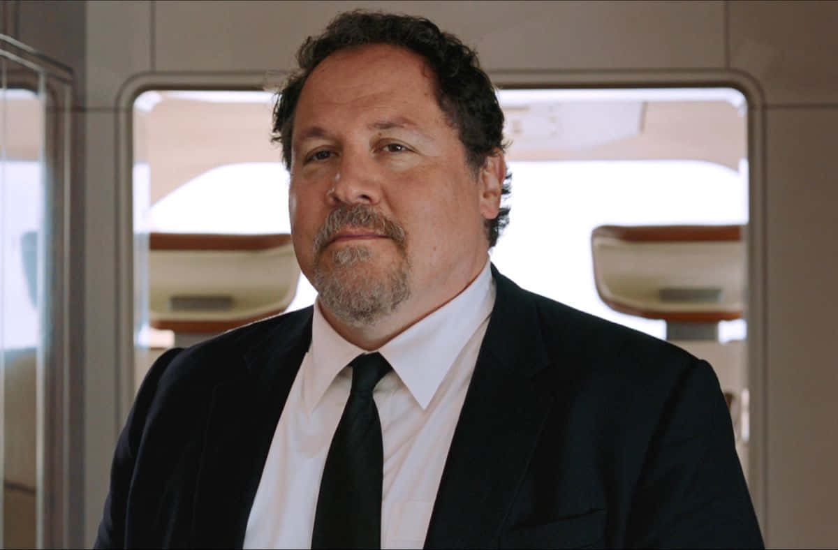 Jon Favreau 桌布 壁紙 — 一名身穿深色西裝及領帶的男子站在飛機機艙內，直視鏡頭，表情嚴肅。