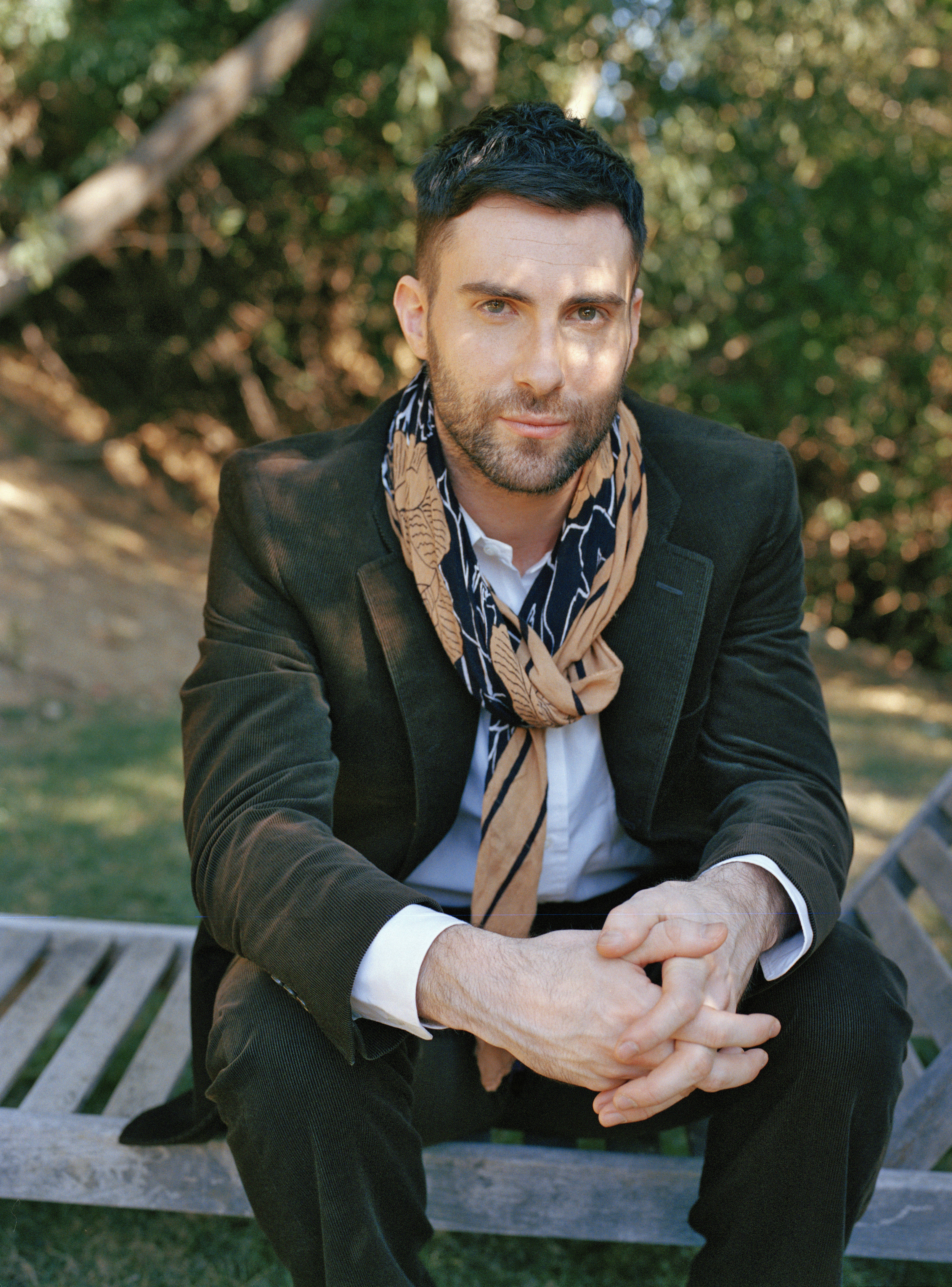 Adam Levine 桌布 壁紙 — 一名身穿深色西裝和圖案圍巾的男子，自信地坐在陽光普照的綠蔭中的木長椅上。
