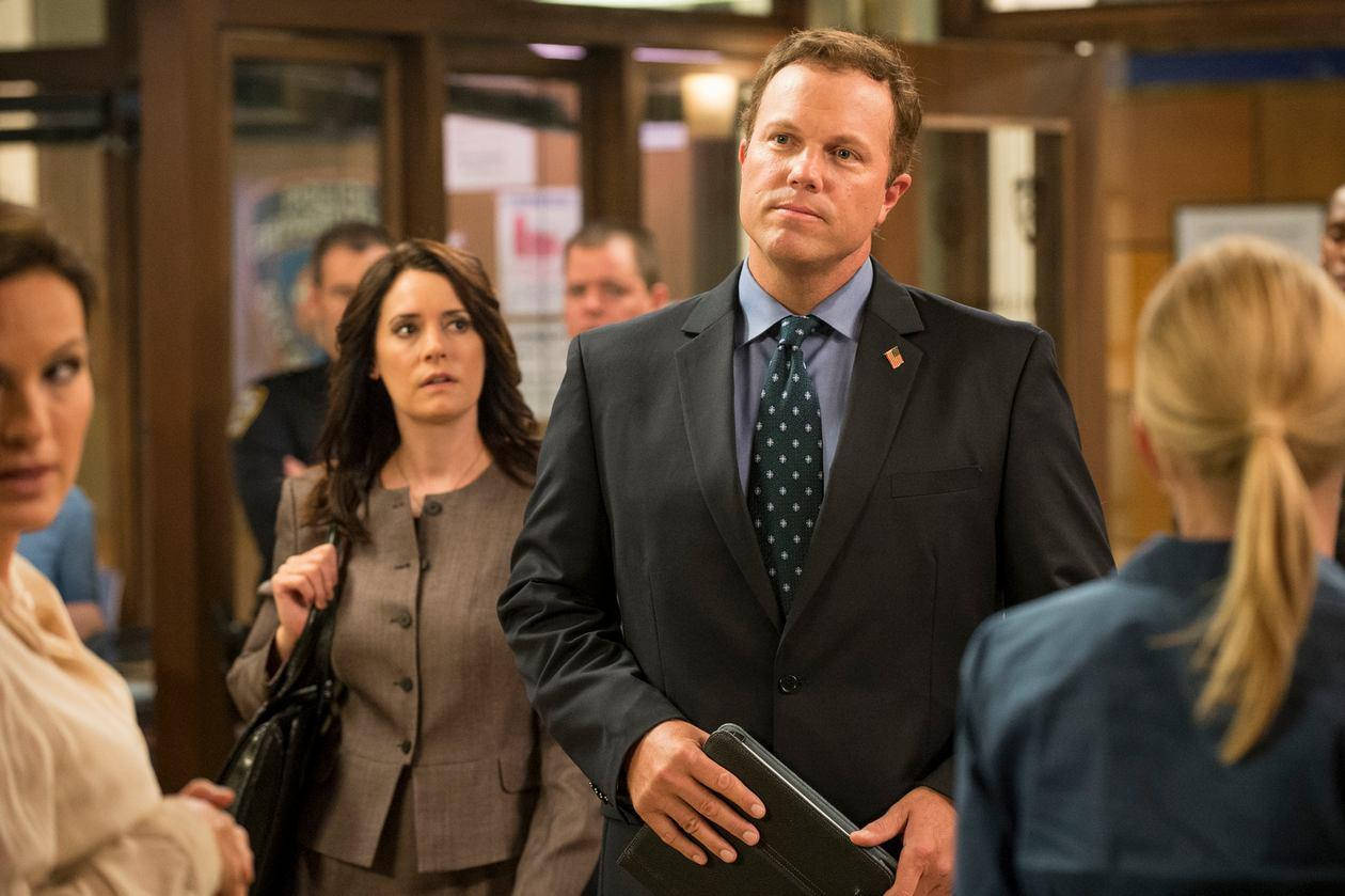 adam baldwin 桌布 壁紙 — 一名身穿深色西裝和圖案領帶的男子站在繁忙的辦公室走廊中，手持文件夾，同事們在旁觀看。