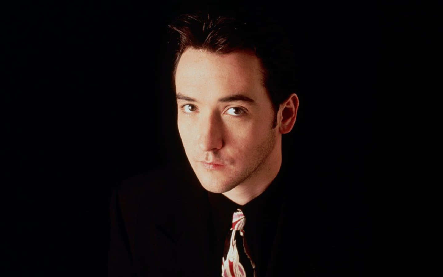 John Cusack 桌布 壁紙 — 一名身穿深色西裝和圖案領帶的男子，在黑色背景和戲劇性燈光下專注地凝視。