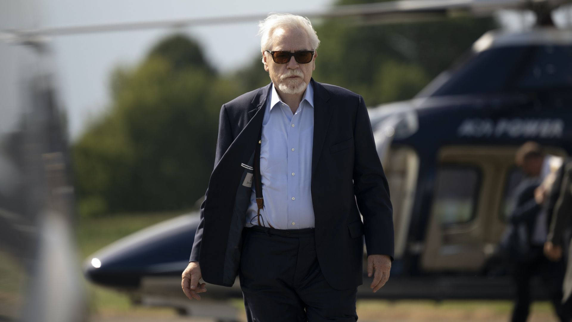 Succession 桌布 壁紙 — 一名身穿深色西裝和太陽眼鏡的男子自信地走向鏡頭，背景中有一架模糊的直升機。