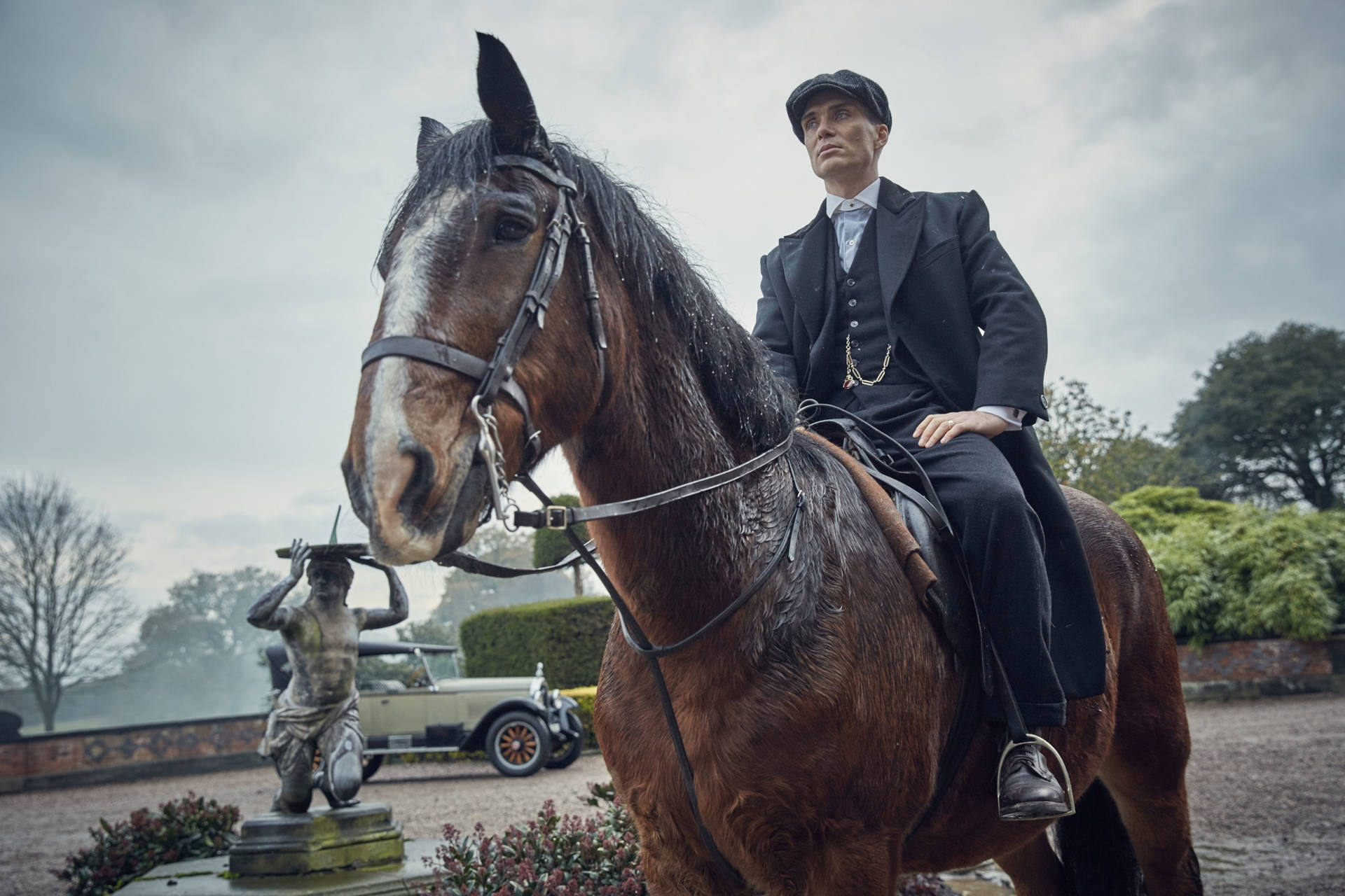 穿西裝的男子在霧氣花園中騎棕色馬，背景有老爺車和雕像。 — Peaky Blinders 壁紙