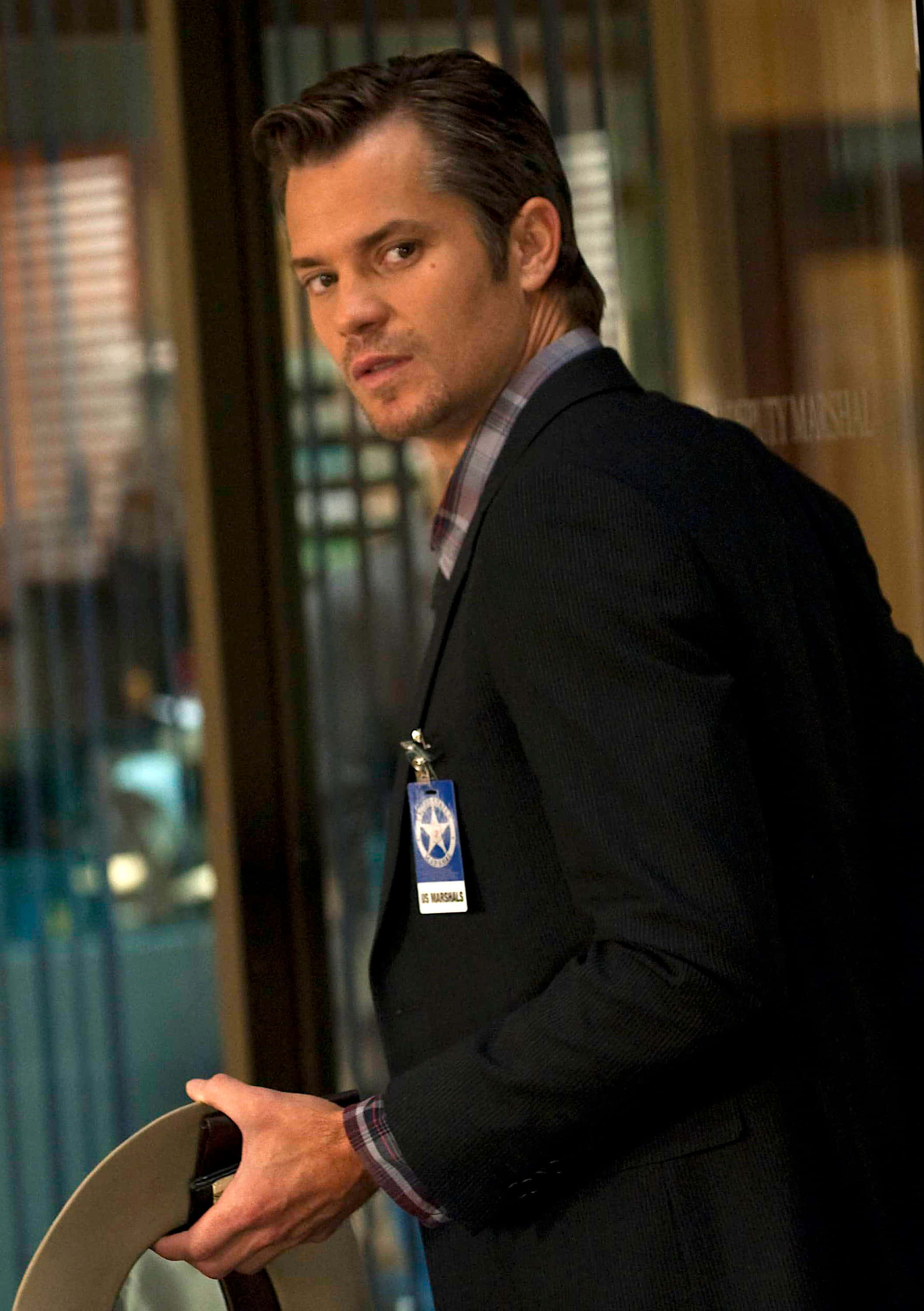 timothy olyphant 桌布 壁紙 — 一名身穿深色西裝和徽章的保全人員站在玻璃門口，手持帽子，神情嚴肅。