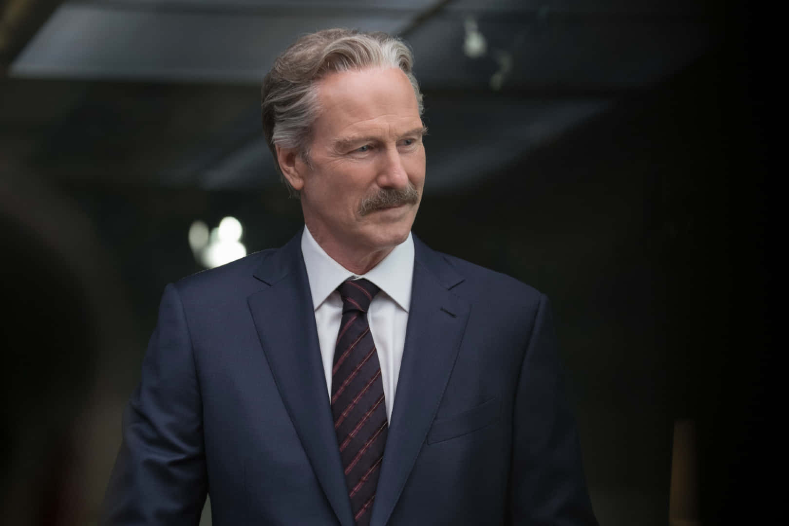 William Hurt 桌布 壁紙 — 一名身穿深色西裝和條紋領帶的男子在昏暗燈光下神情嚴肅地站立。