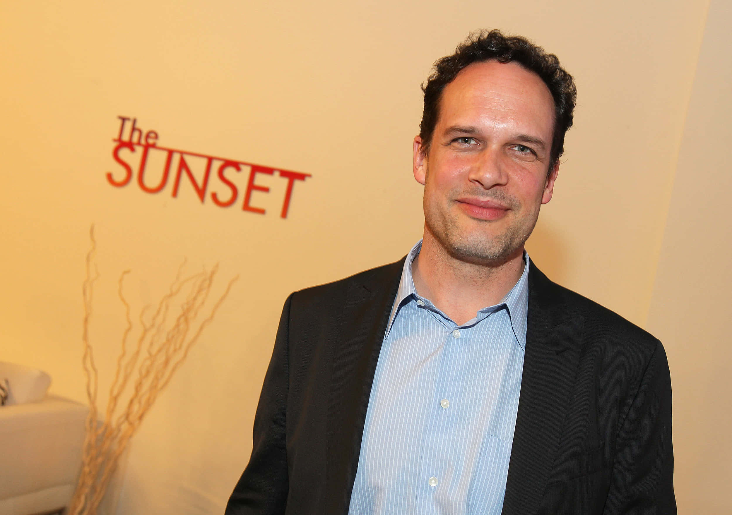 Diedrich Bader 桌布 壁紙 — 一名身穿深色西裝和淺藍色襯衫的男子在米色牆壁前對著鏡頭微笑，牆上有紅色的「The SUNSET」文字和乾燥的樹枝。