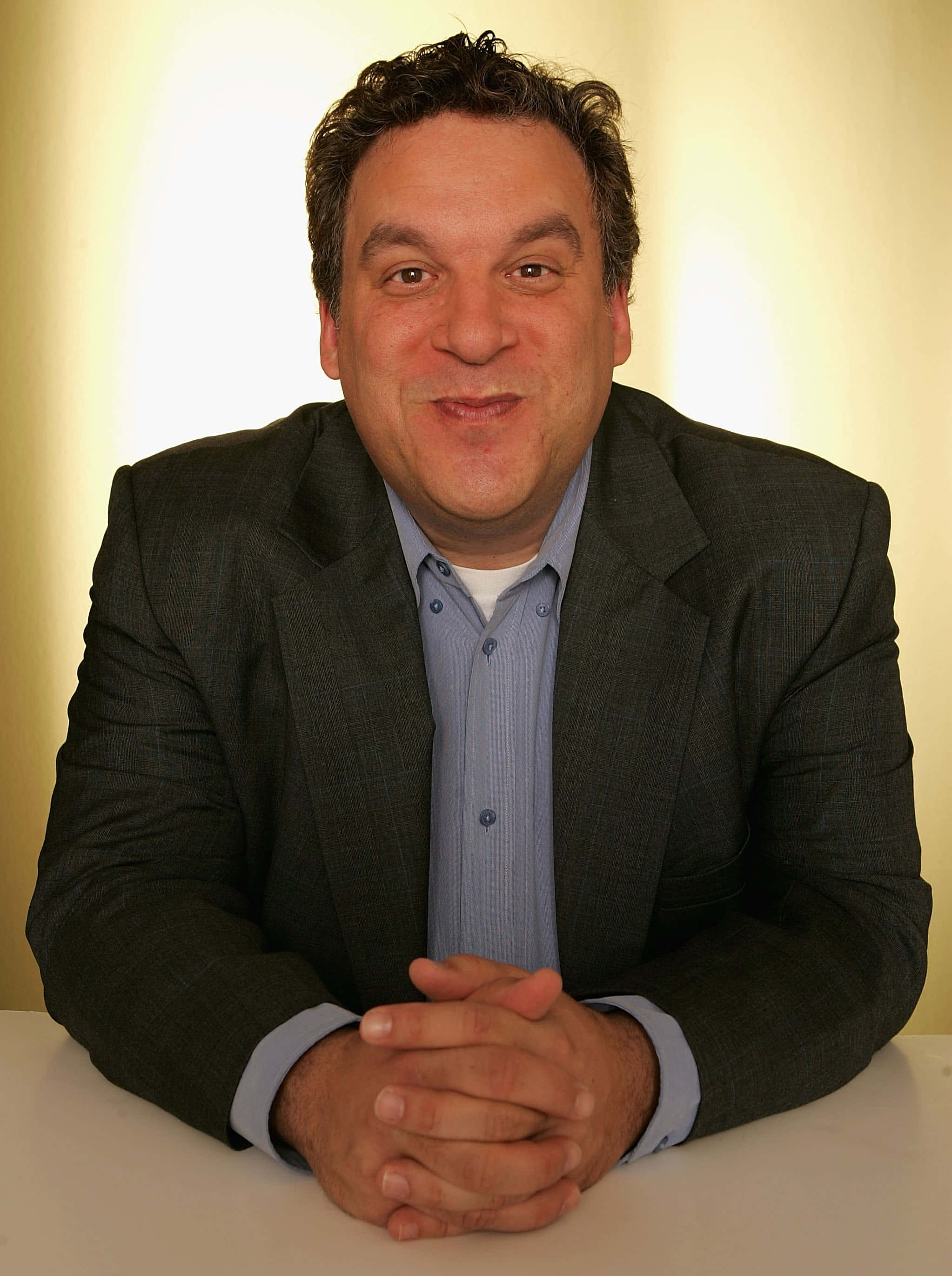 Jeff Garlin 桌布 壁紙 — 一名身穿深色西裝和淺藍色襯衫的男子，在柔和的黃色背景前坐在白色桌子旁，露出溫暖的笑容。