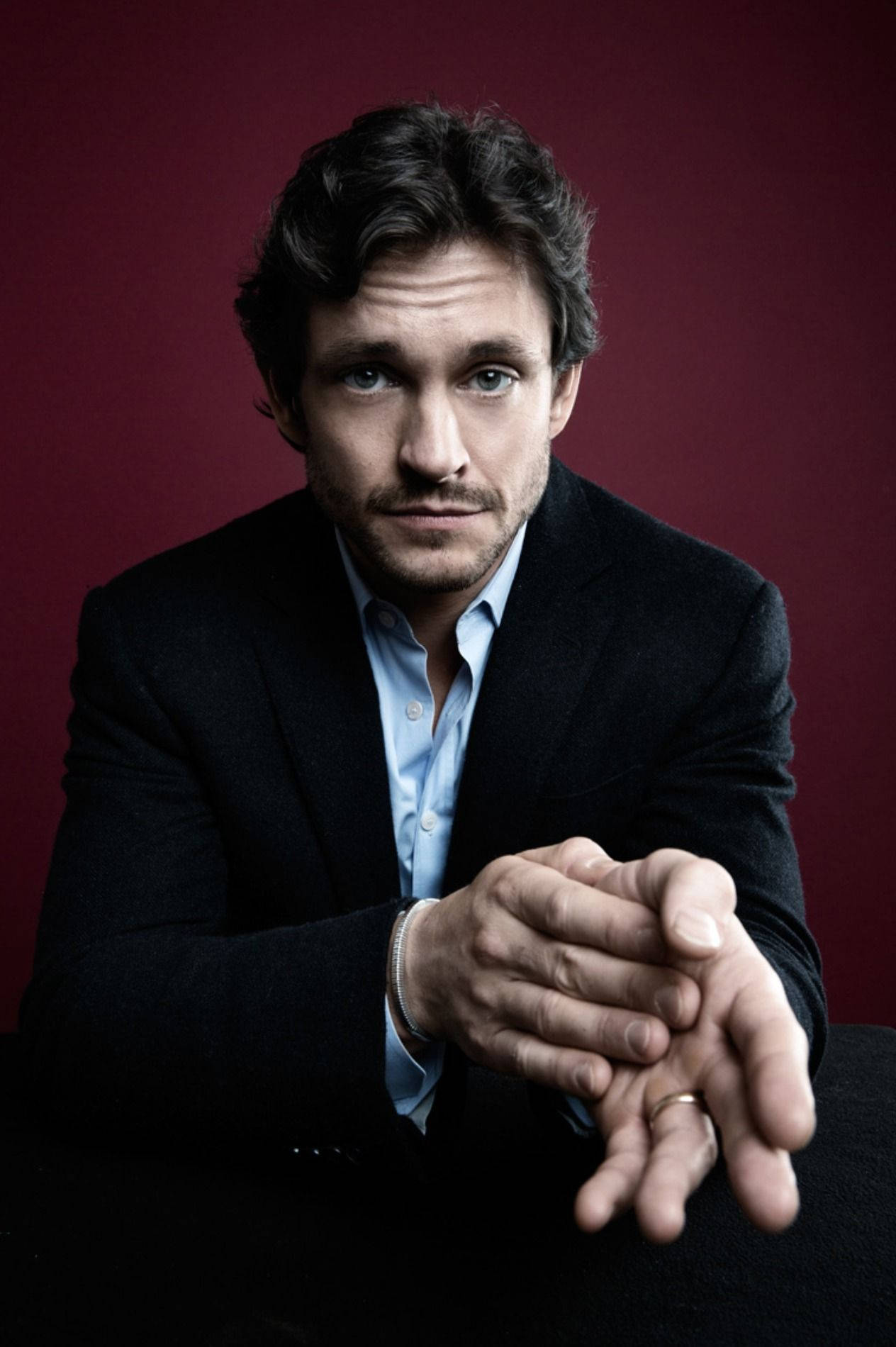 Hugh Dancy 桌布 壁紙 — 一名身穿深色西裝和淺藍色襯衫的男子，雙手緊握，神情嚴肅，坐在深紅色背景前。