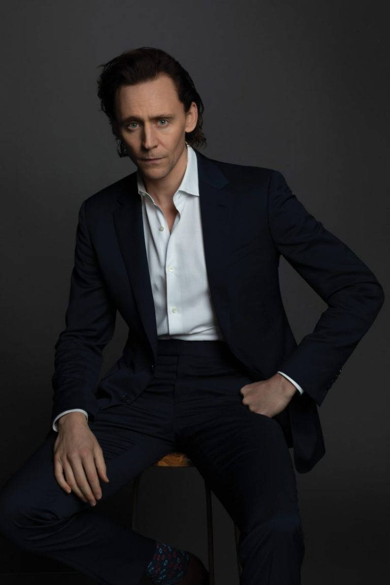 Tom 桌布 壁紙 — 一名身穿深色西裝和白色襯衫的男子自信地坐在灰色背景前的凳子上，眼神專注。