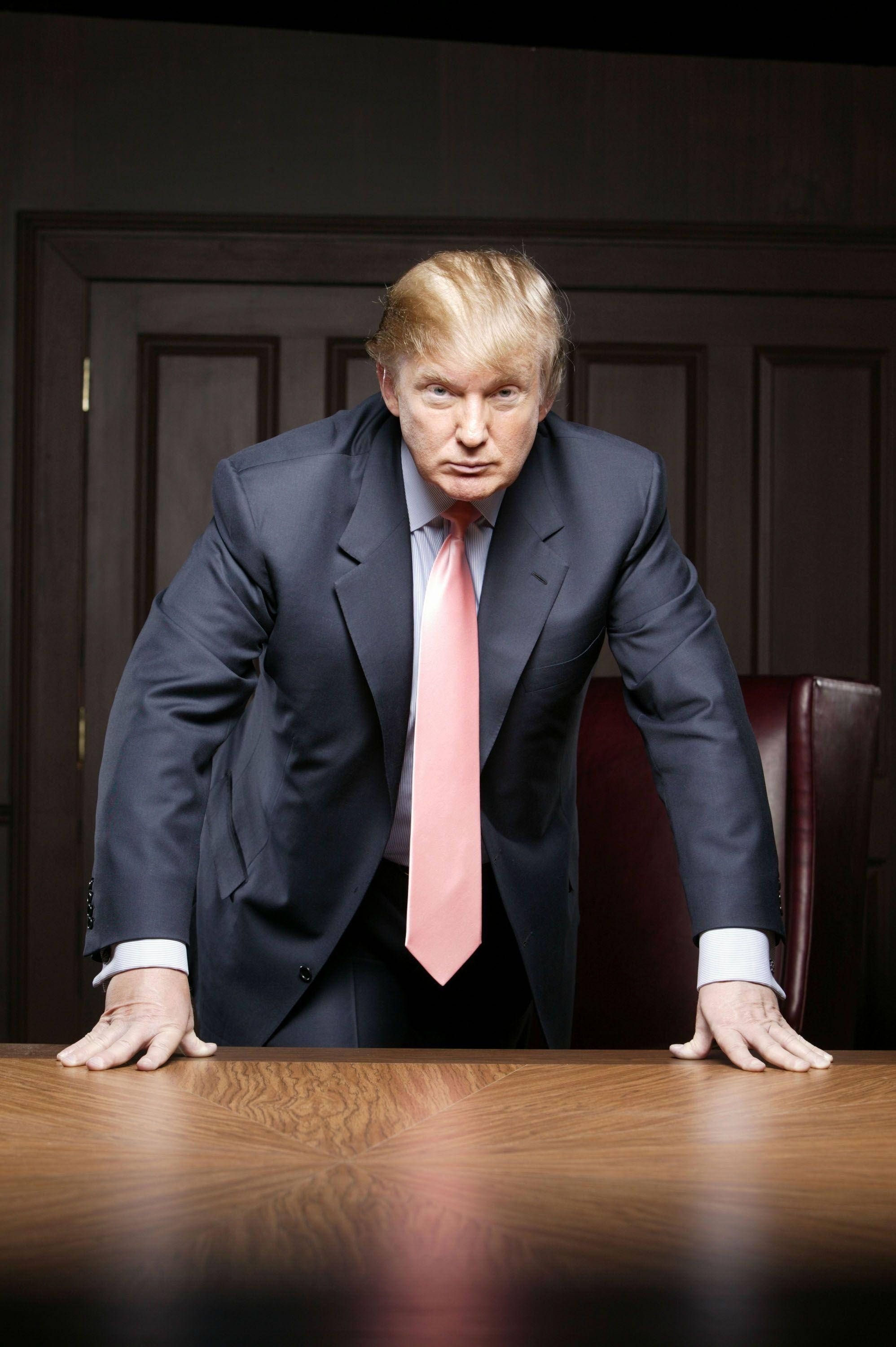 donald trump 桌布 壁紙 — 一名身穿深色西裝和粉紅色領帶的男子在燈光昏暗的辦公室裡，帶著強烈的表情俯身在一張木桌上。
