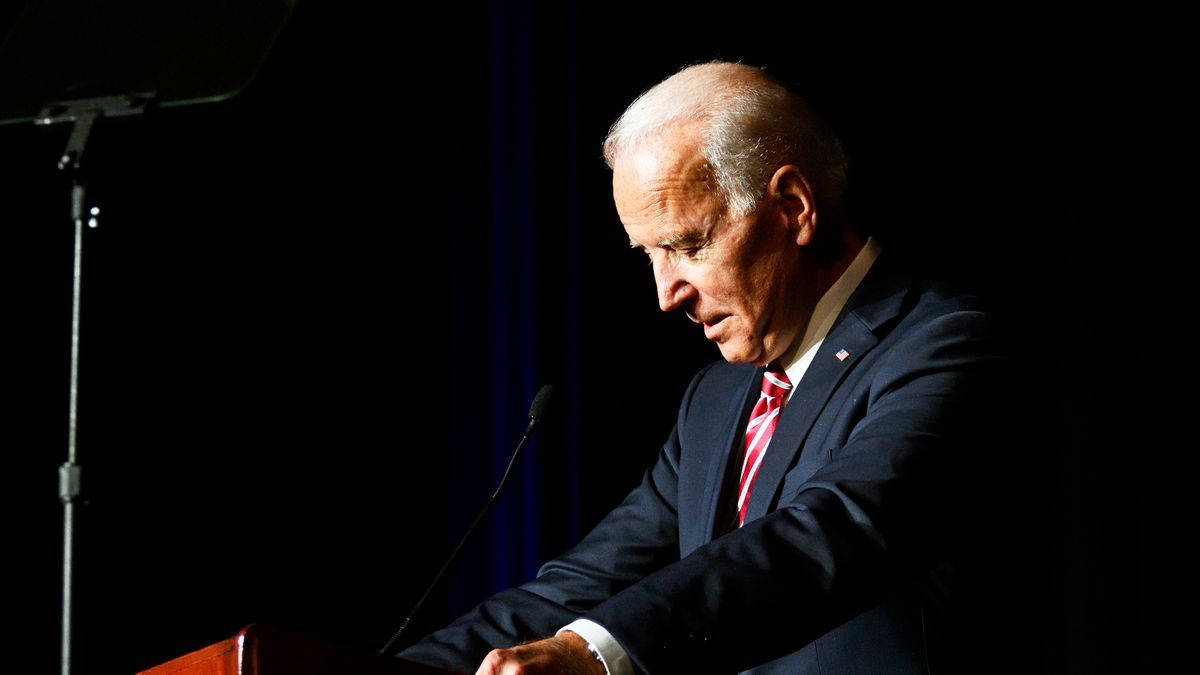 Biden 桌布 壁紙 — 一名身穿深色西裝和紅色領帶的男子站在木製講台上，在黑色背景下受到戲劇性的聚光燈照射。
