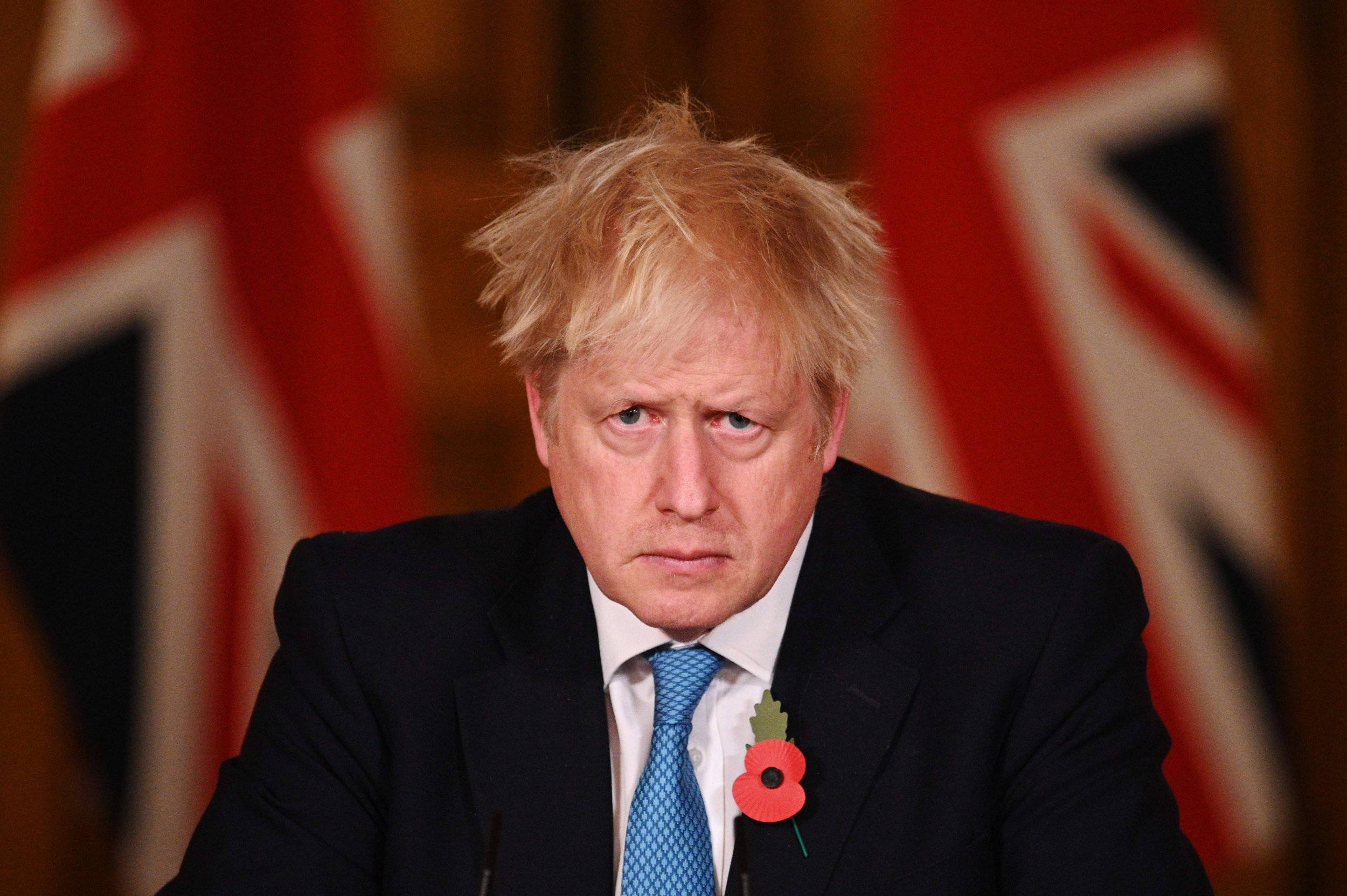 Boris Johnson 桌布 壁紙 — 一名身穿深色西裝和藍色領帶的男子，在模糊的英國國旗背景下佩戴紅色罌粟花胸針。