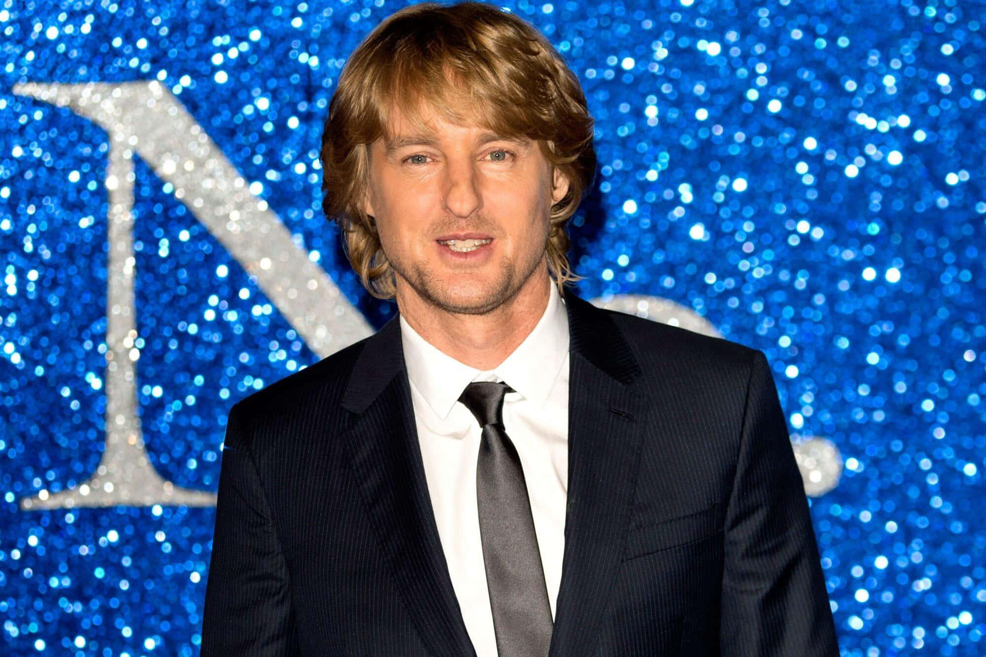 Owen Wilson 桌布 壁紙 — 一名身穿深色西裝和領帶的男子，在閃閃發光的藍色亮片背景前微笑，背景上有一個銀色字母。