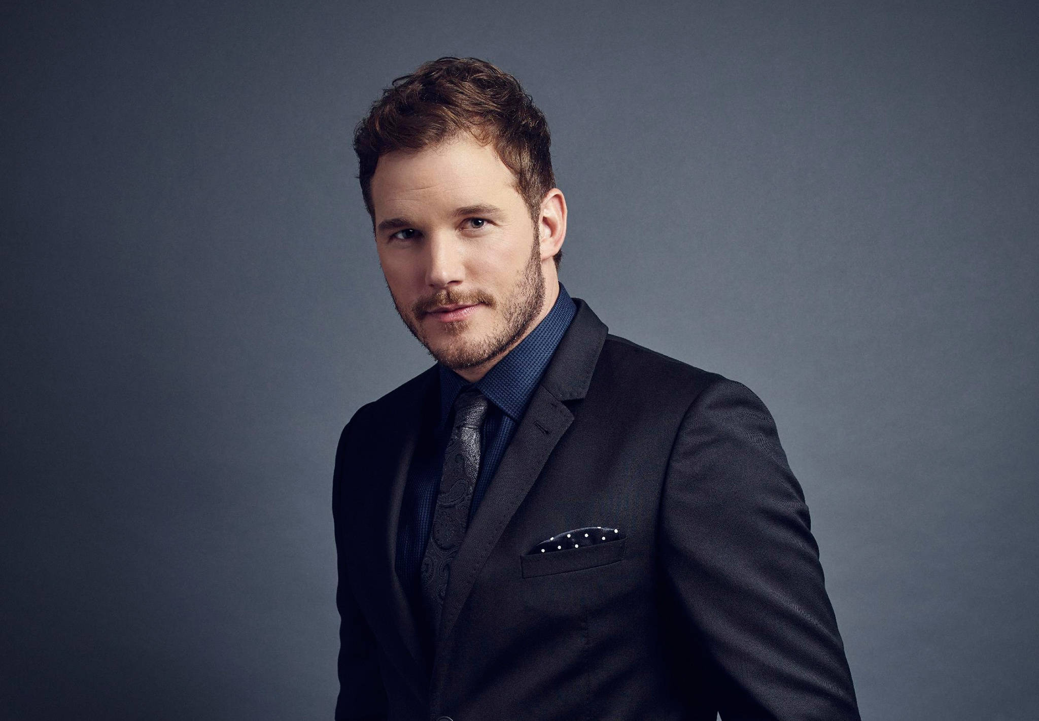Chris Pratt 桌布 壁紙 — 一名身穿深色西裝和領帶的男子在柔和燈光的灰色攝影棚背景下自信地擺姿勢。