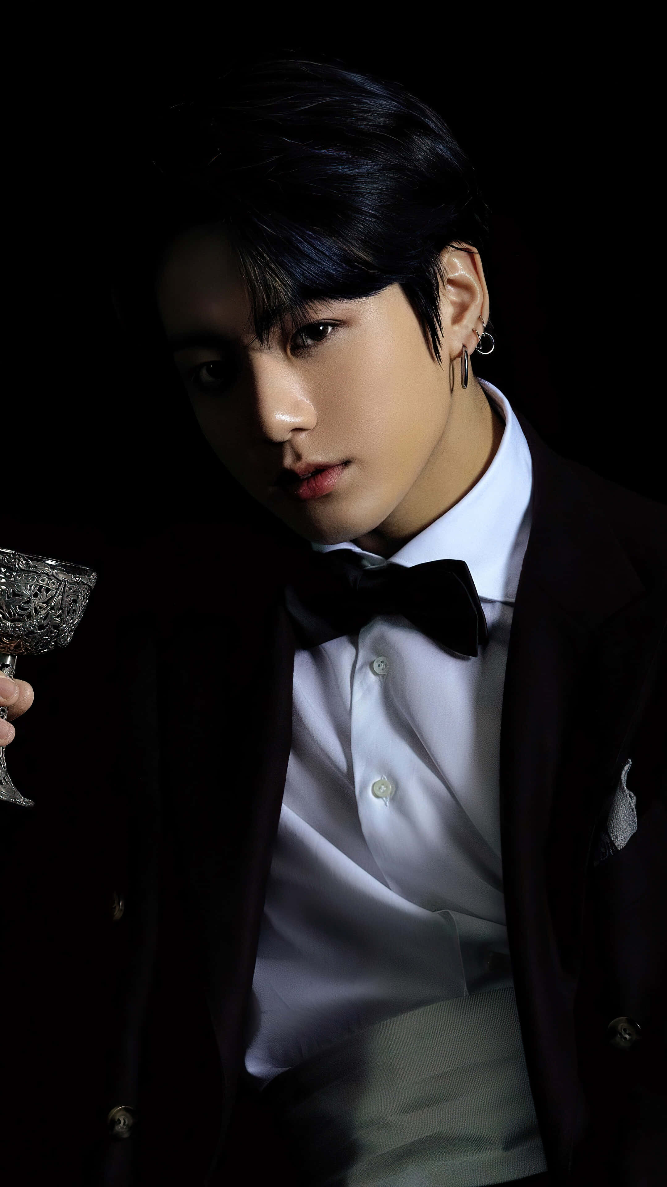 Jungkook 桌布 壁紙 — 一名身穿深色西裝和領結的男子，在黑色背景和戲劇性燈光下拿著一個華麗的玻璃杯。