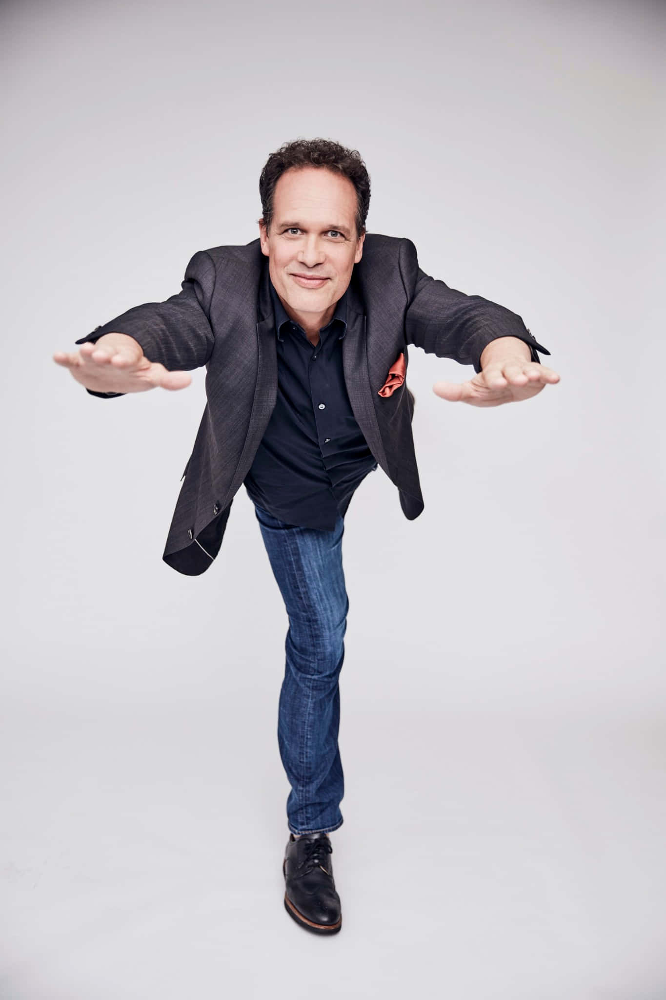 Diedrich Bader 桌布 壁紙 — 一名身穿深色西裝外套和牛仔褲的男子，在純白色攝影棚背景前張開雙臂擺出俏皮的姿勢。