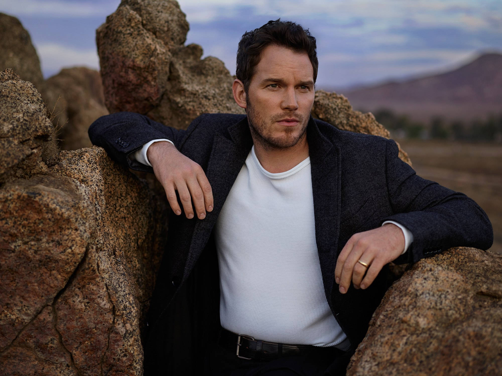 Chris Pratt 桌布 壁紙 — 一名身穿深色西裝外套和白色襯衫的男子，在多雲的天空下，倚靠在沙漠景觀中的崎嶇岩石上。