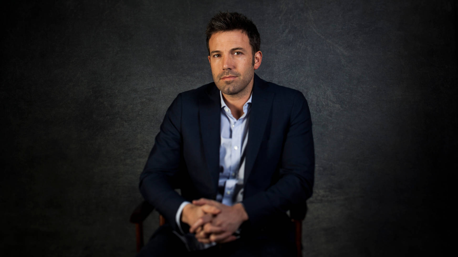 Ben Affleck 桌布 壁紙 — 一名身穿深色西裝的男子，在柔和的攝影棚燈光下，平靜地坐在有紋理的灰色背景前。