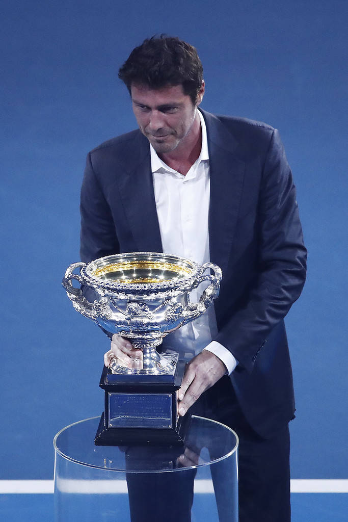 Tennis 桌布 壁紙 — 一名身穿深色西裝的男子，在藍色網球場背景下，輕輕地拿著一個放在玻璃底座上的大型華麗銀色獎盃。