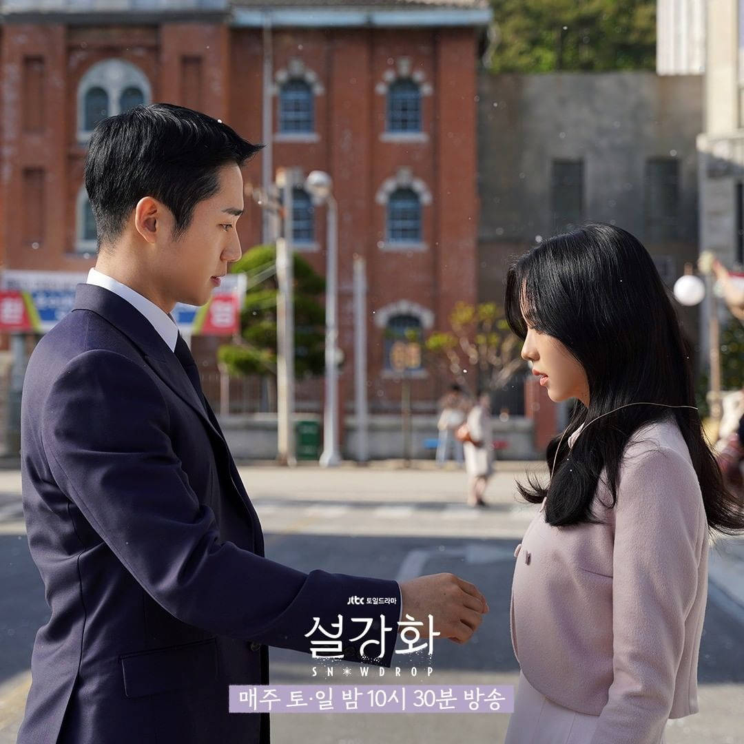 drama 桌布 壁紙 — 一名身穿深色西裝的男子和一名身穿淺粉色外套的女子在陽光明媚的城市街道上相對而立，身後是一棟紅磚建築。
