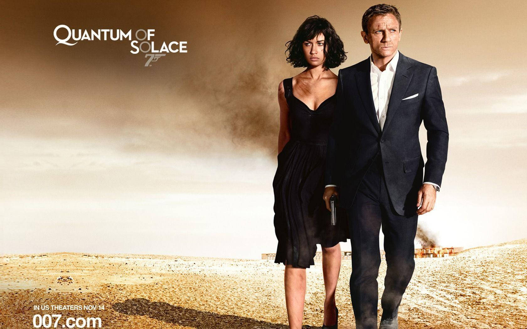 Bond 桌布 壁紙 — 一名身穿深色西裝的男子和一名身穿黑色連衣裙的女子站在荒蕪的沙漠中，天空朦朧，身後煙霧升騰。