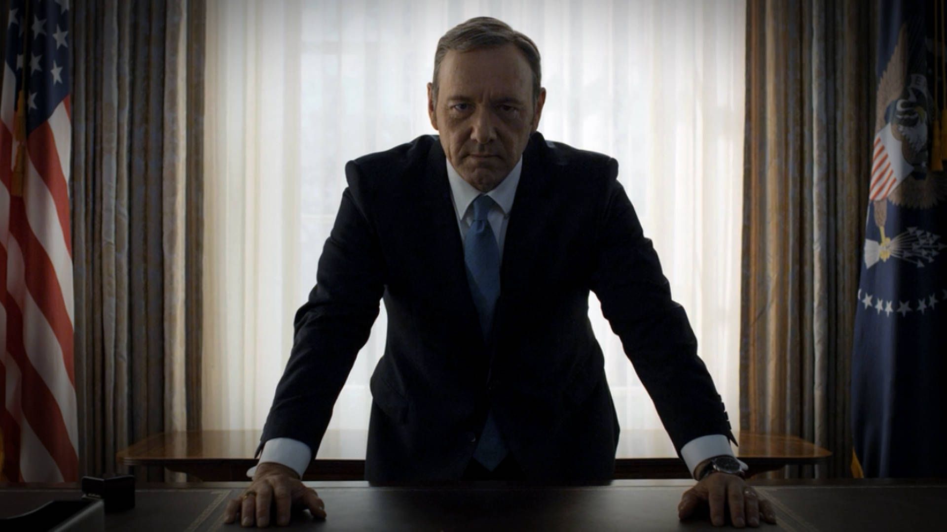 house of cards 桌布 壁紙 — 一名身穿深色西裝的男子在一個官方辦公室裡，身後有美國國旗，身體前傾靠在桌子上。