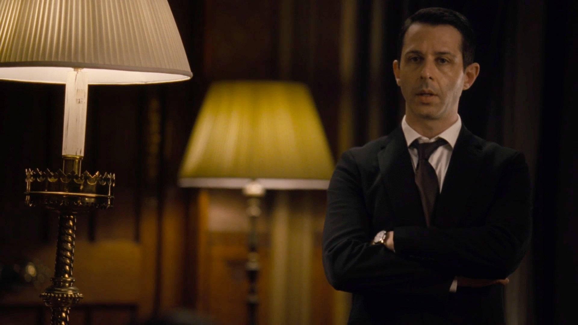 Succession 桌布 壁紙 — 一名身穿深色西裝的男子在一個溫暖照明的房間裡交叉雙臂站立，房間內有華麗的燈具散發出柔和的金色光芒。