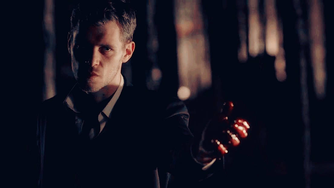 Vampire Diaries 桌布 壁紙 — 一名身穿深色西裝的男子在夜間陰暗的森林中，手持一個發光的紅色物體，眼神專注。
