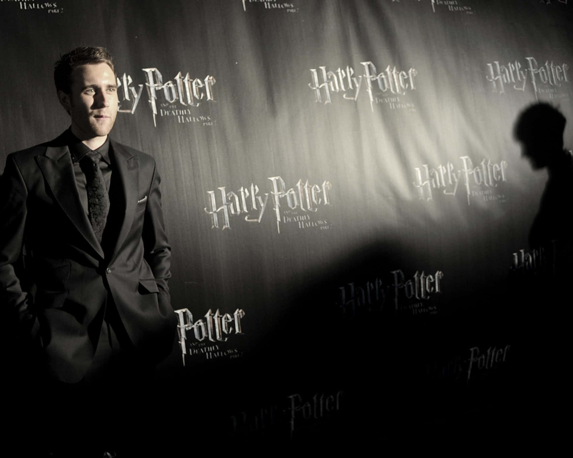 Neville Longbottom 桌布 壁紙 — 一名身穿深色西裝的男子在戲劇性的聚光燈下自信地站在哈利波特電影背景前。