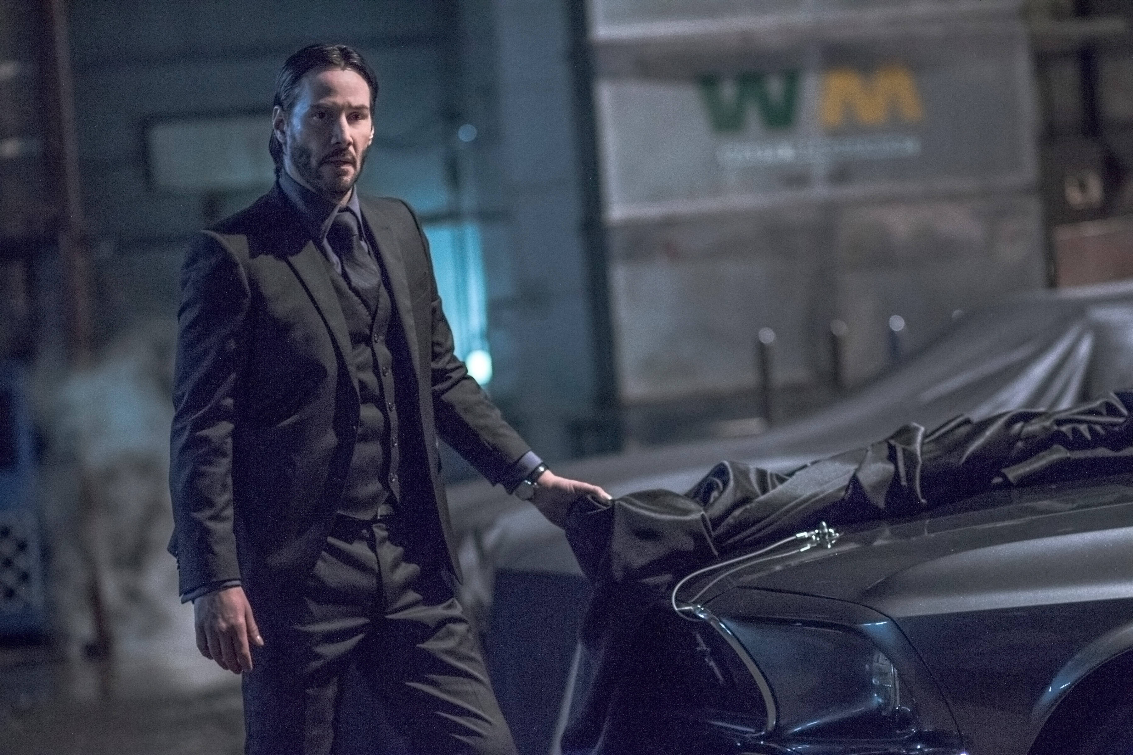 John Wick 桌布 壁紙 — 一名身穿深色西裝的男子在晚上站在一輛時尚的汽車旁邊，引擎蓋上有一具被覆蓋的屍體，身處工業環境中。