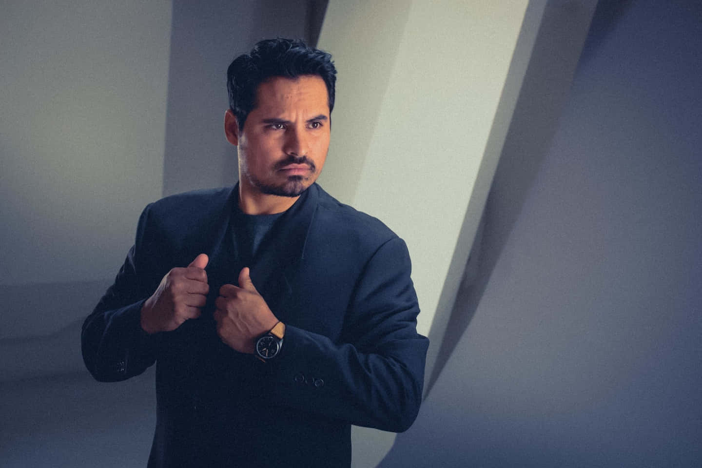 Michael Peña 桌布 壁紙 — 一名身穿深色西裝的男子在極簡幾何牆壁前，在戲劇性燈光下調整他的外套。