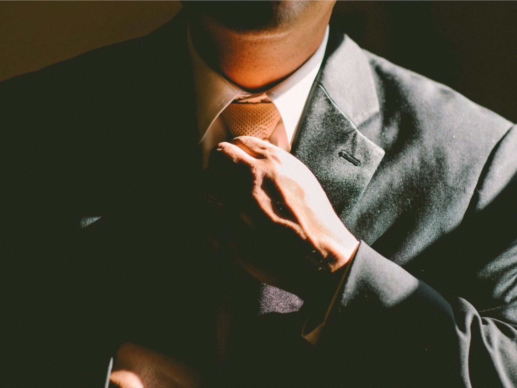 商業 桌布 壁紙 — 一名身穿深色西裝的男子在溫暖的方向性燈光下調整他的圖案領帶，投射出戲劇性的陰影。
