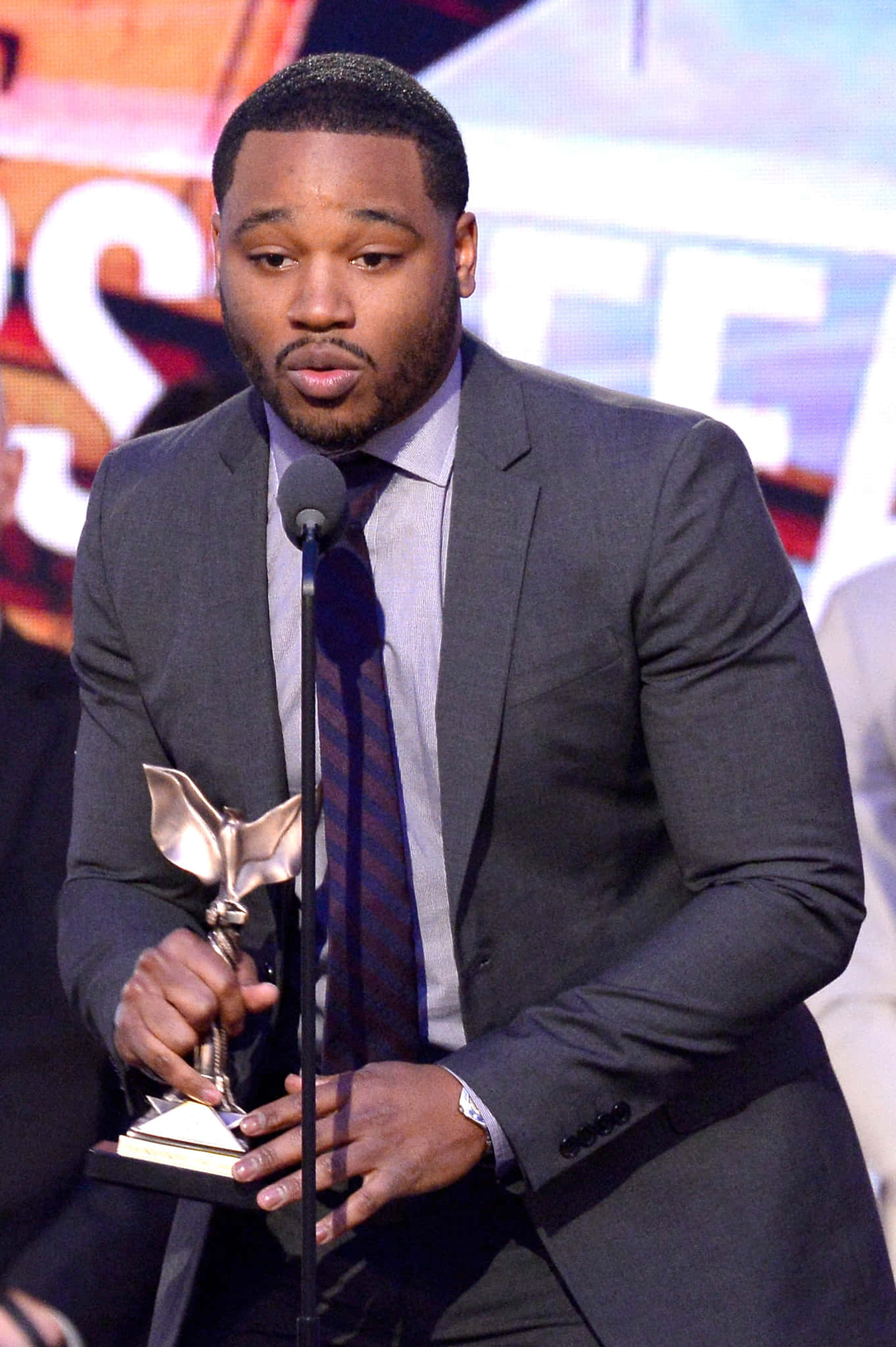 Ryan Coogler 桌布 壁紙 — 一名身穿深色西裝的男子在燈光明亮的舞台活動中手持金屬翼狀獎項。