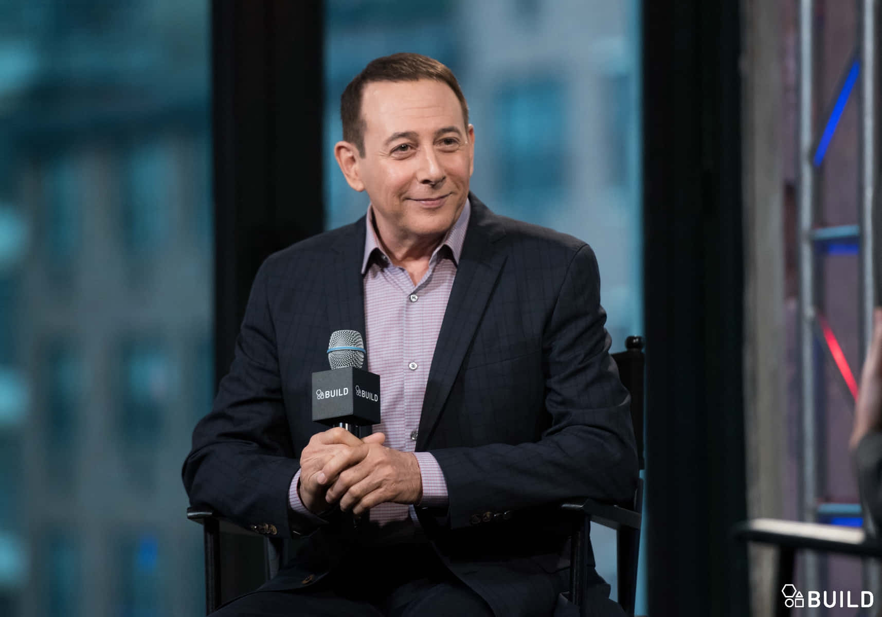 Paul Reubens 桌布 壁紙 — 一名身穿深色西裝的男子在現代化攝影棚內接受採訪，手持麥克風微笑，背景是模糊的城市天際線。