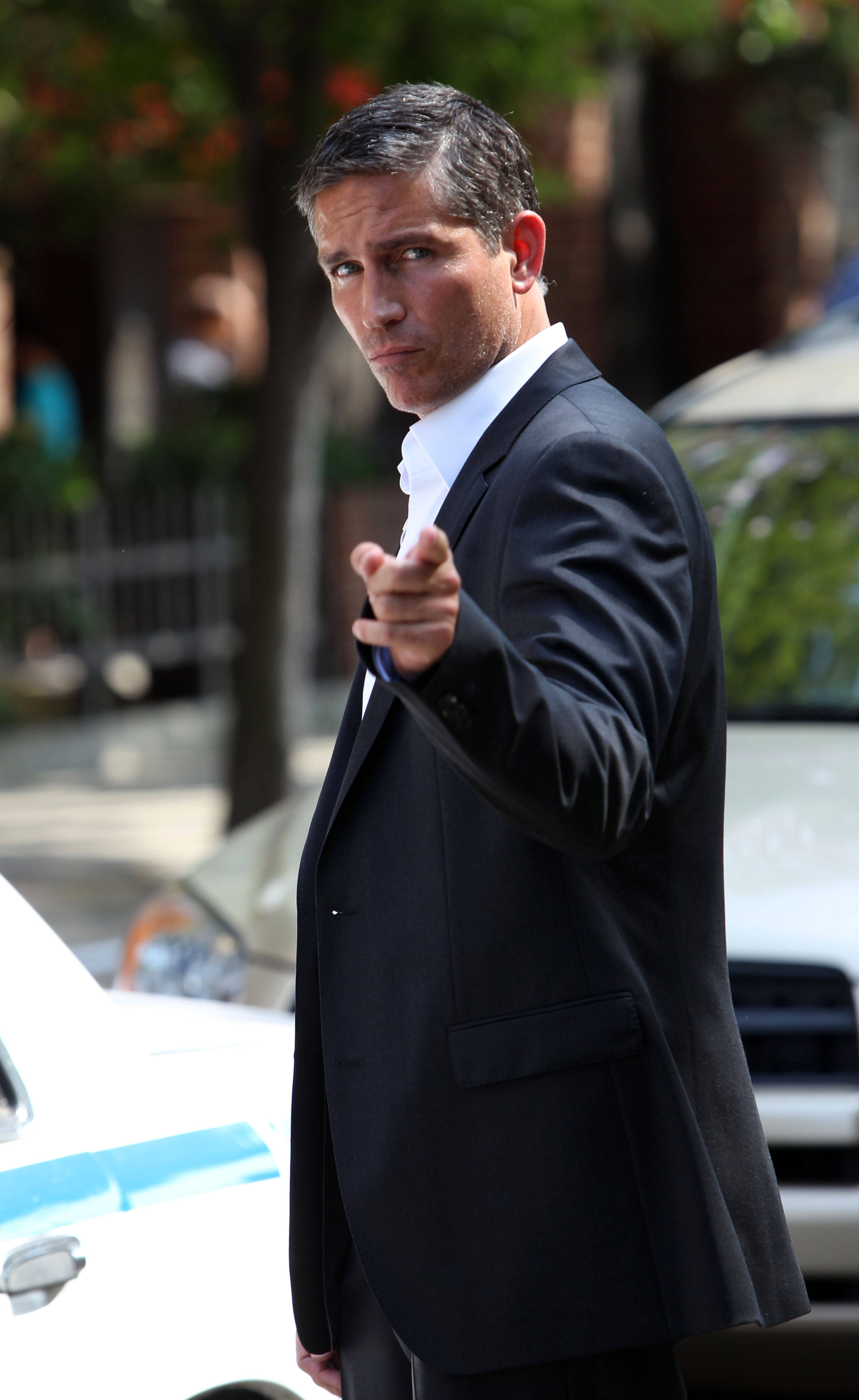 actor 桌布 壁紙 — 一名身穿深色西裝的男子在陽光明媚的城市街道上，直接指向觀看者，街道上有停泊的汽車和樹木。