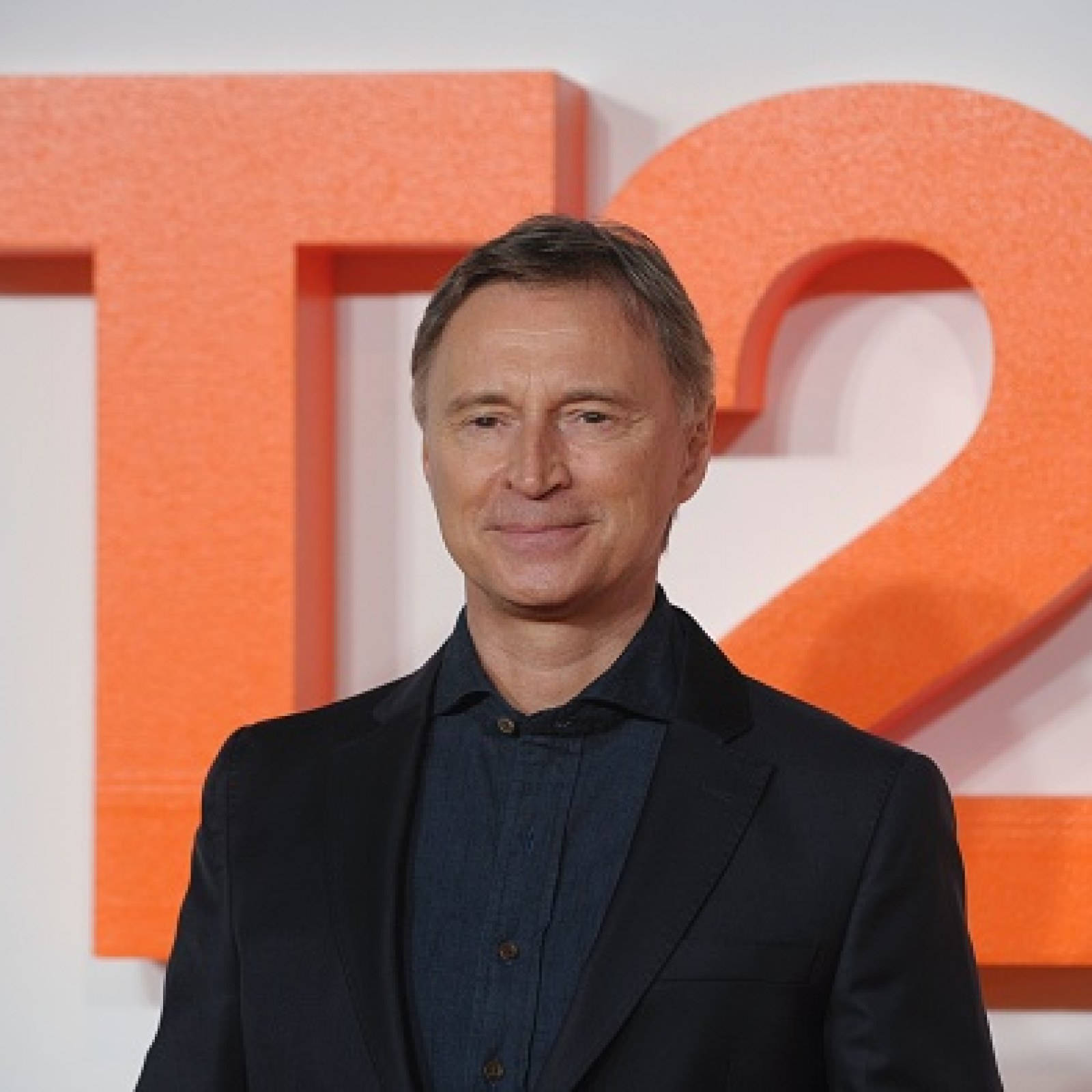 Smile 桌布 壁紙 — 一名身穿深色西裝的男子在鮮明的橙色「T2」背景下溫柔微笑。