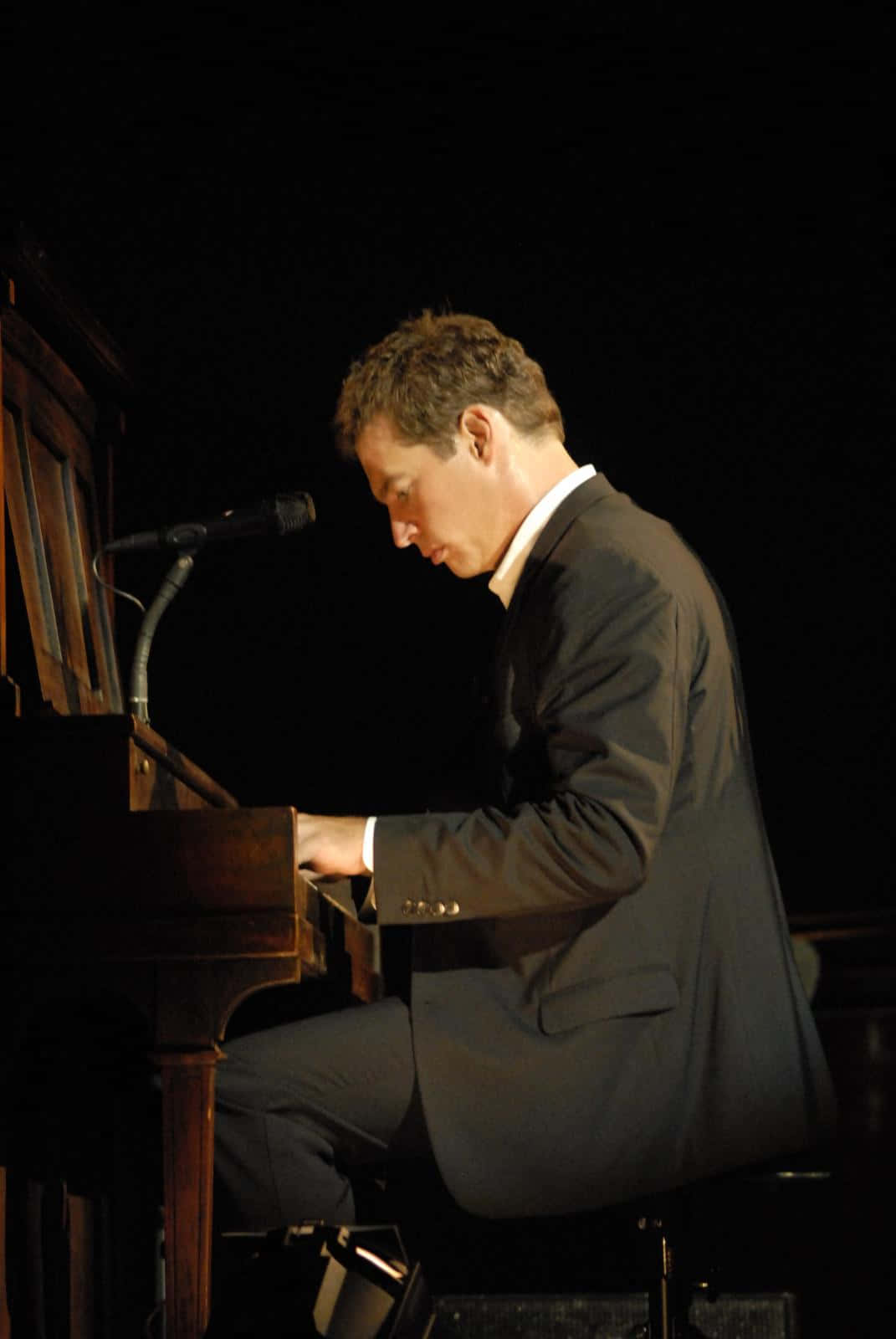 Harry Connick Jr 桌布 壁紙 — 一名身穿深色西裝的男子在黑色背景的舞台燈光下彈奏三角鋼琴，附近有一個麥克風。