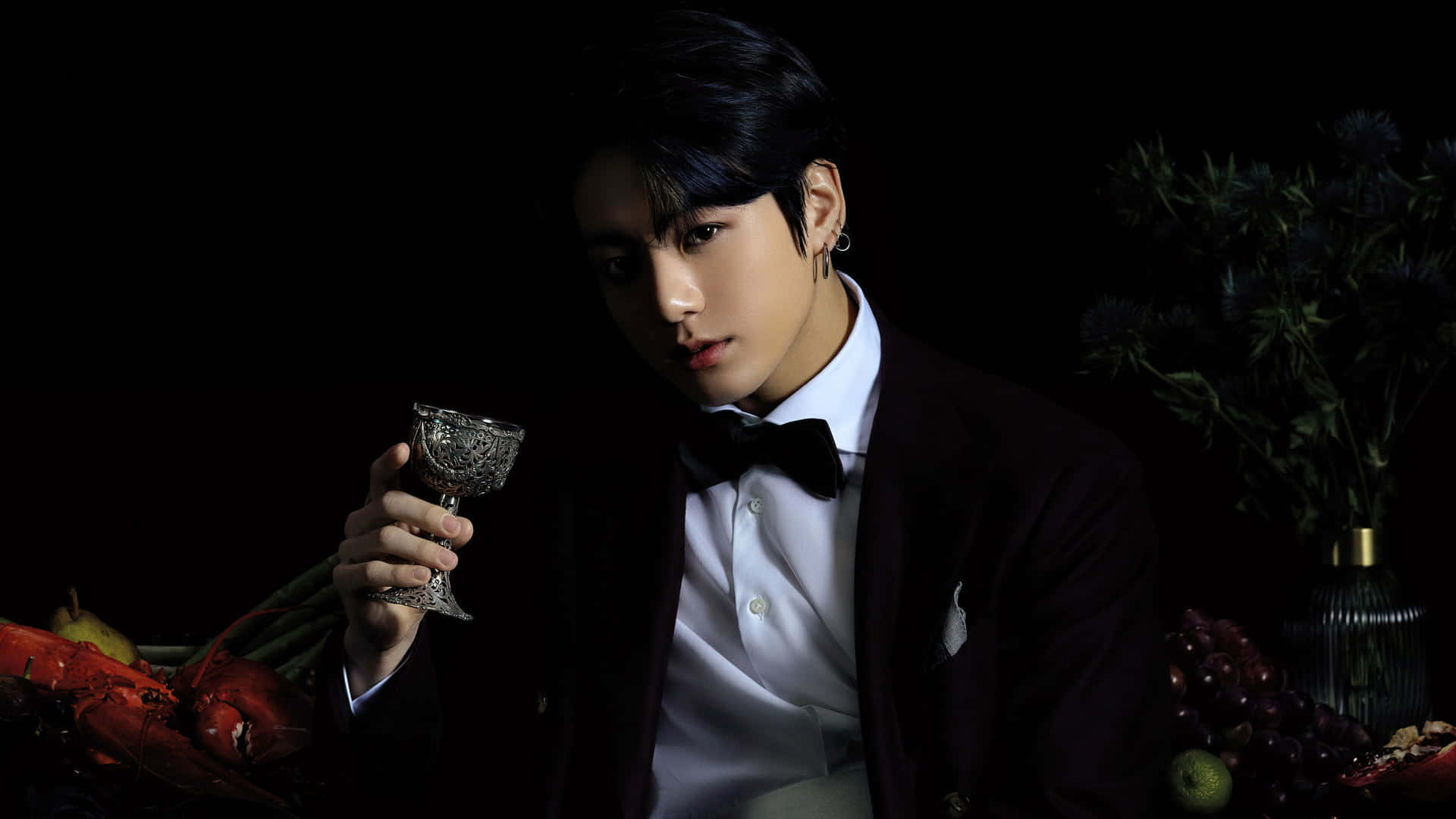 Jungkook 桌布 壁紙 — 一名身穿深色西裝的男子在黑色背景前拿著一個華麗的玻璃杯，背景有紅色水果和綠色植物。