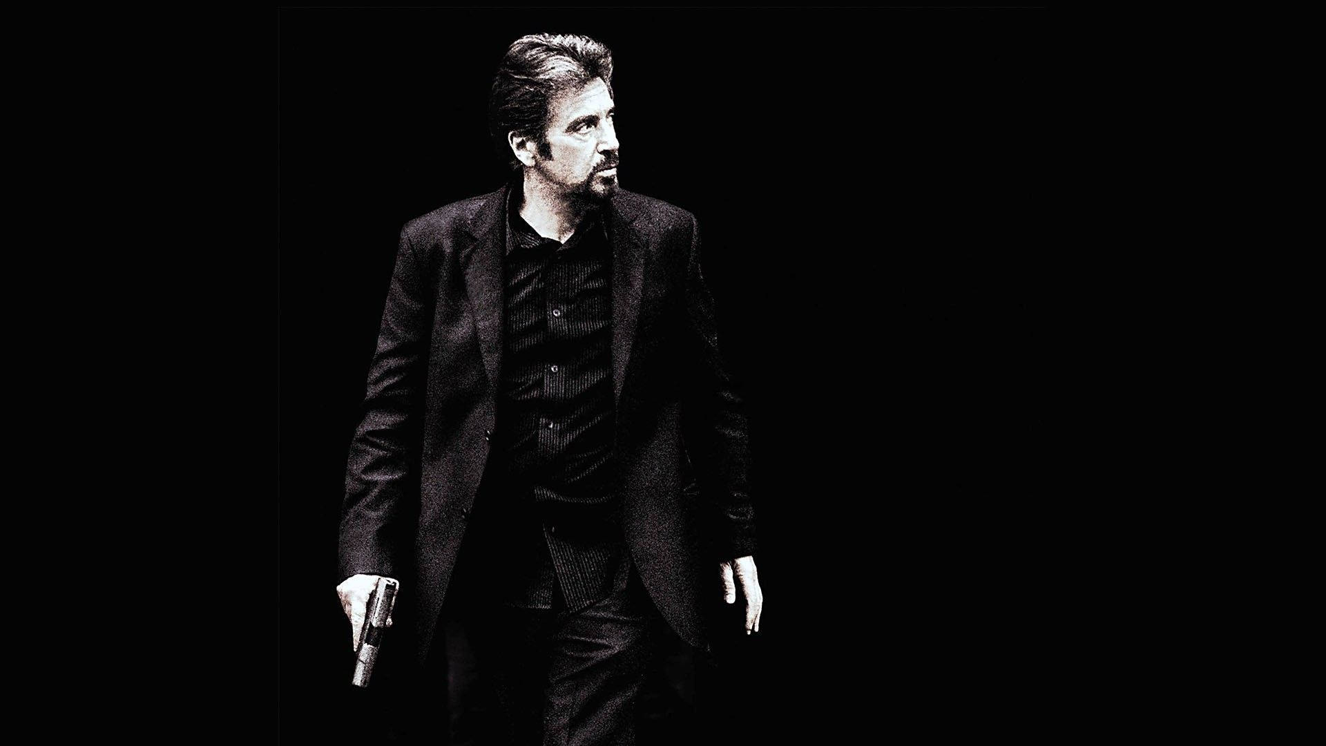 Al Pacino 桌布 壁紙 — 一名身穿深色西裝的男子在黑色背景和戲劇性燈光下行走時手持槍支。
