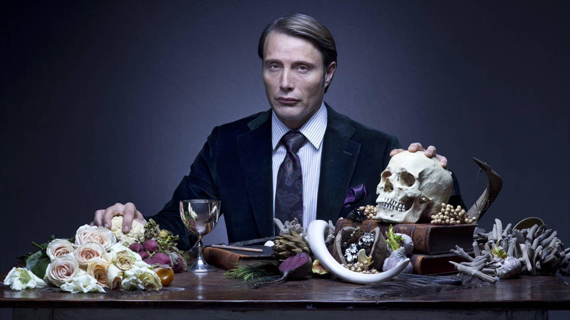 Hannibal 桌布 壁紙 — 一名身穿深色西裝的男子坐在一張被頭骨、鮮花和鹿角環繞的桌子旁，在戲劇性的工作室燈光下。