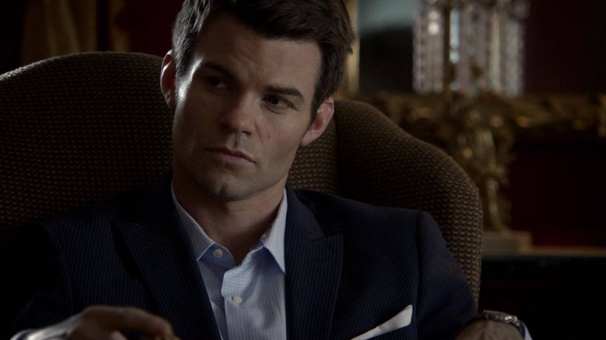 elijah mikaelson 桌布 壁紙 — 一名身穿深色西裝的男子坐在一張豪華椅子上，表情嚴肅，身處燈光昏暗的華麗房間。