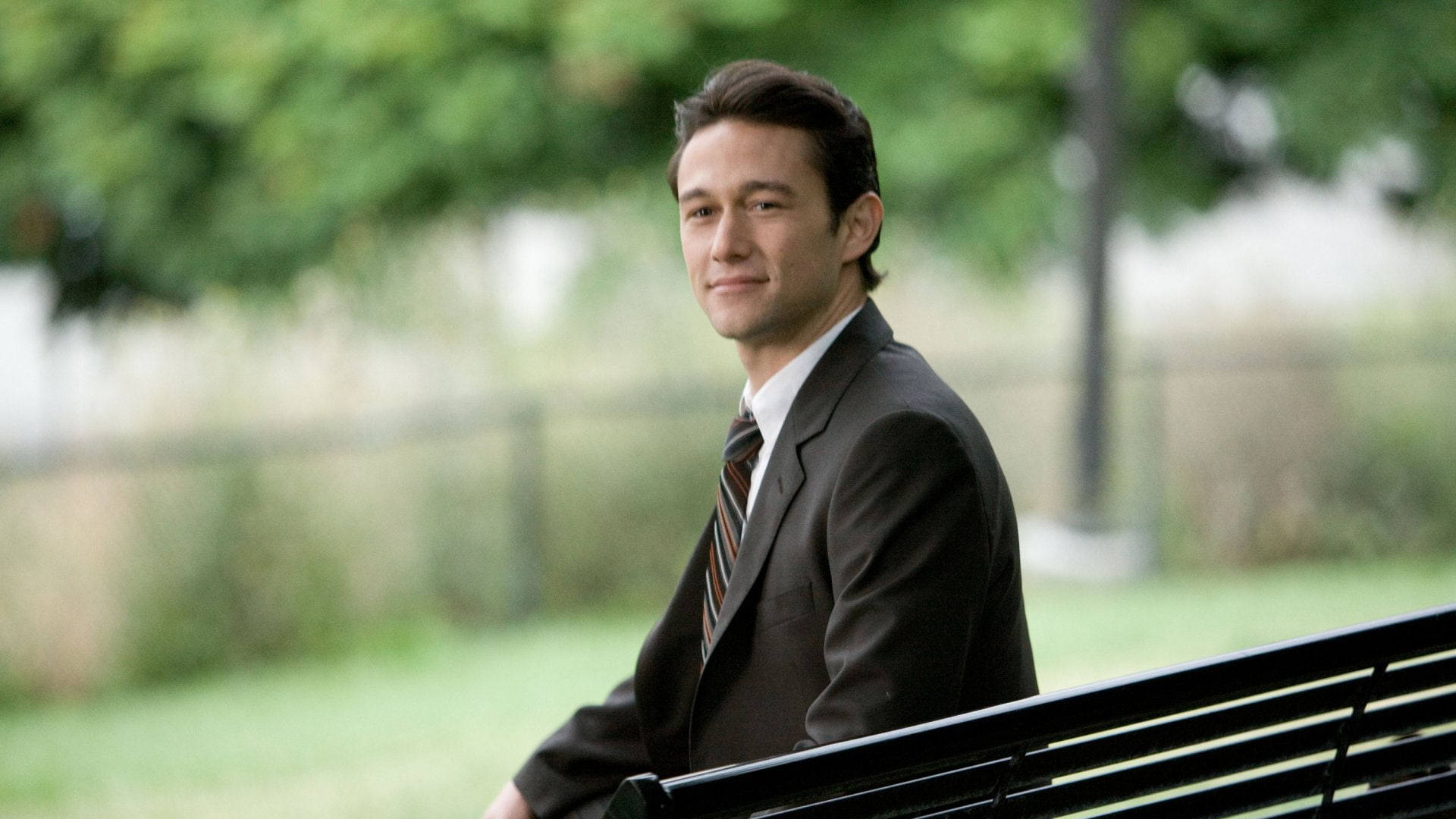 500 Days Of Summer 桌布 壁紙 — 一名身穿深色西裝的男子坐在公園長椅上，帶著溫柔的微笑，周圍是模糊的綠植和柔和的白晝光線。