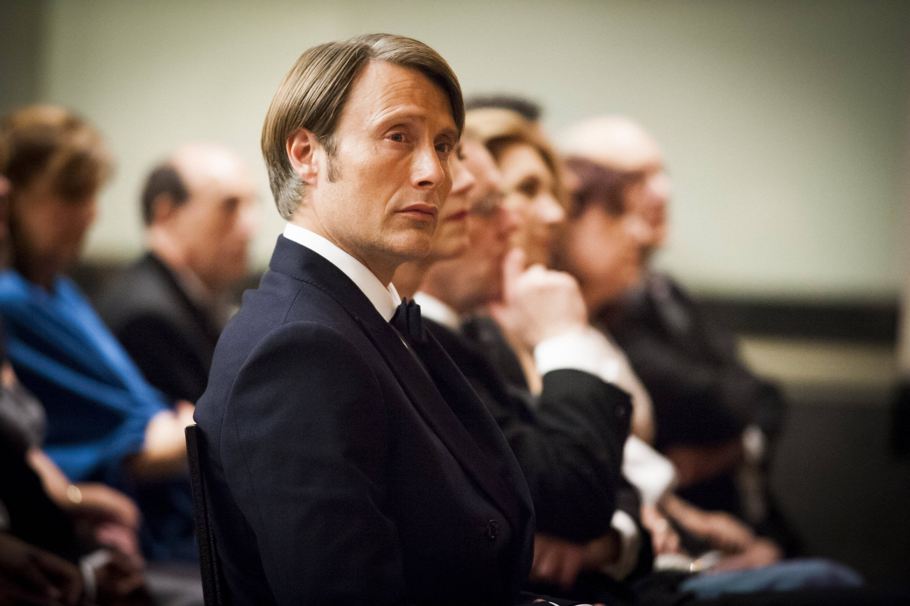 Mads Mikkelsen 桌布 壁紙 — 一名身穿深色西裝的男子坐在燈光昏暗的禮堂中，表情嚴肅，背景中其他人模糊不清。