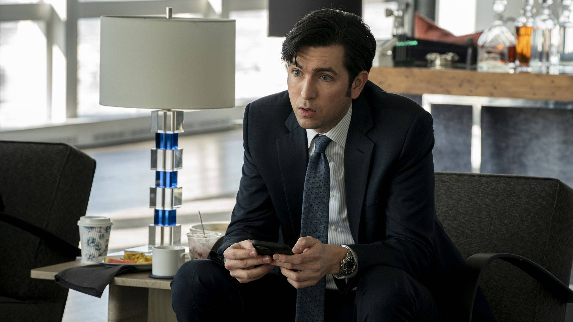 Succession 桌布 壁紙 — 一名身穿深色西裝的男子坐在現代休息室，震驚地看着手機，附近有燈和飲料。