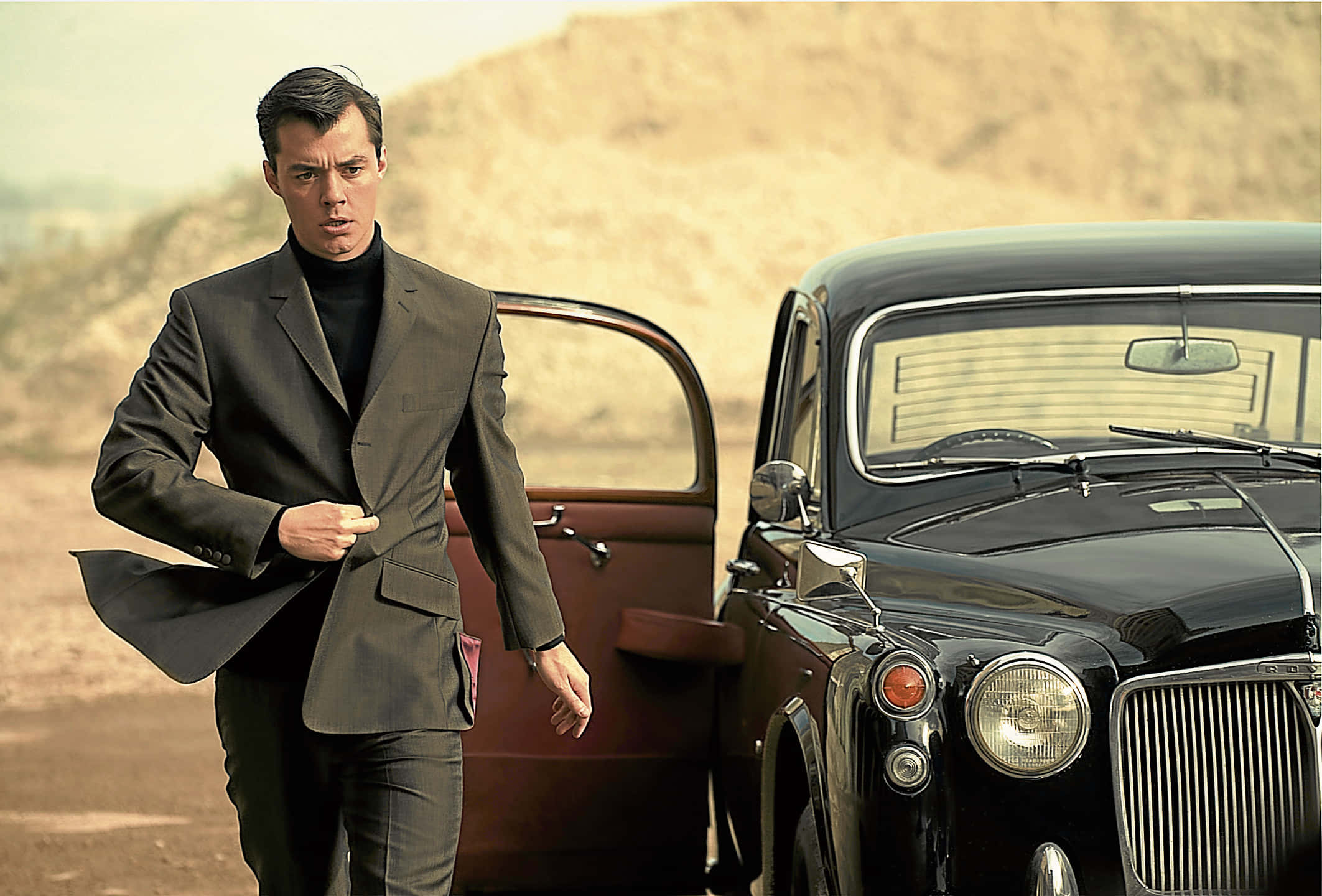 Alfred Pennyworth 桌布 壁紙 — 一名身穿深色西裝的男子從一輛停在塵土飛揚的沙漠景觀中的復古黑色汽車中走出，天色朦朧。