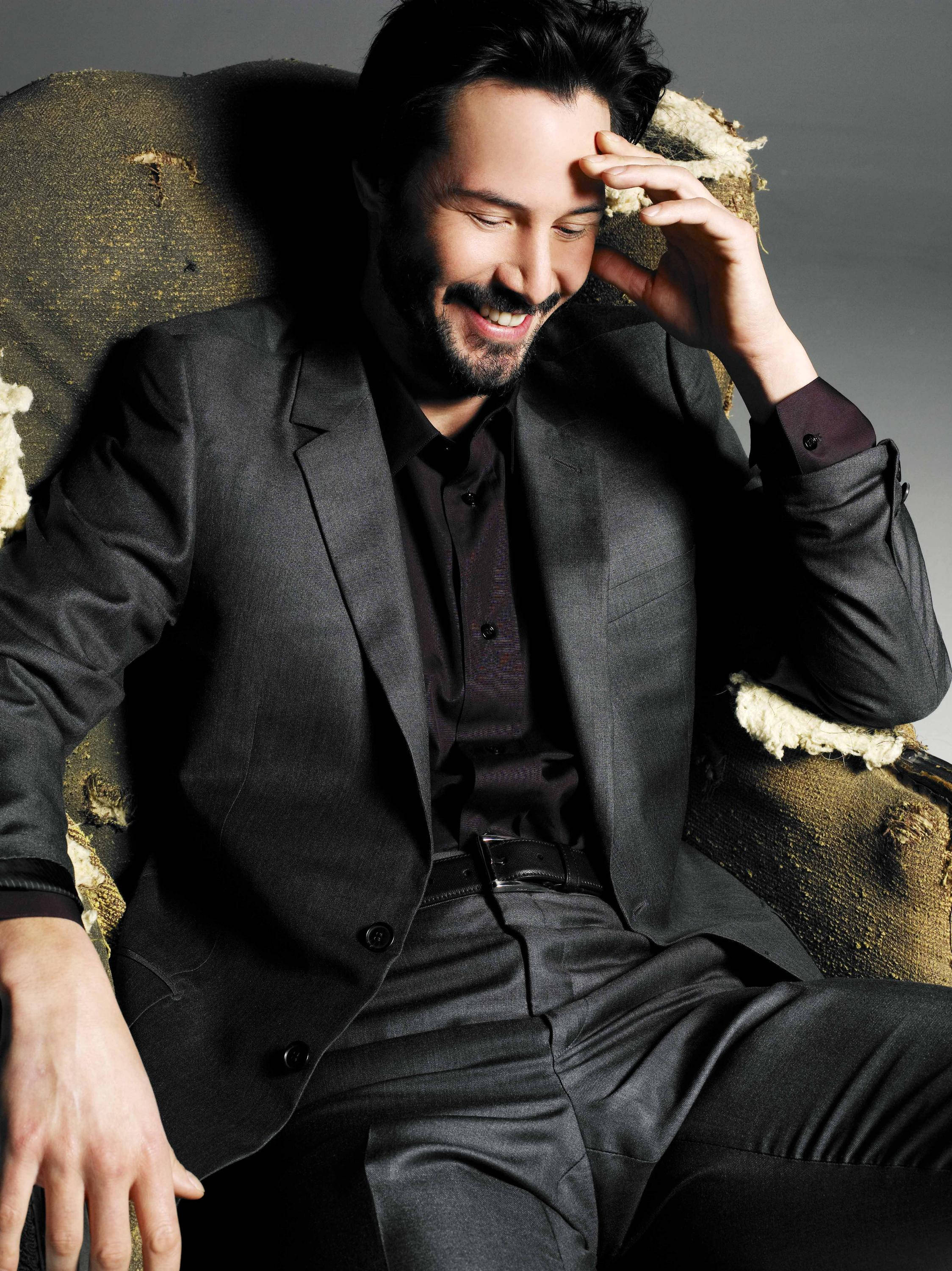 Keanu 桌布 壁紙 — 一名身穿深色西裝的男子微笑著坐在一張磨損的紋理扶手椅上，一隻手放在額頭上。